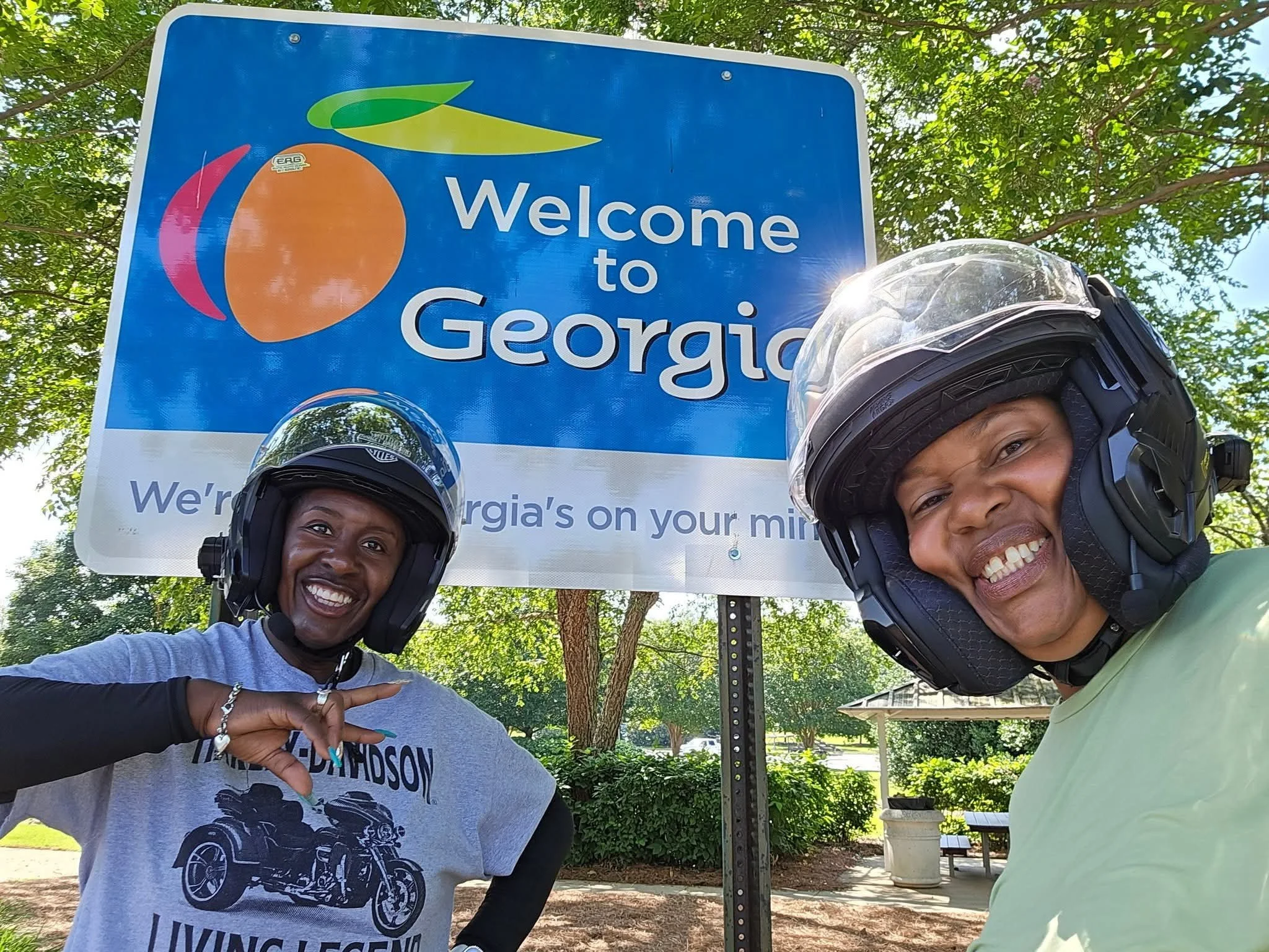 Two women wearing motorcycle helmets standing in front of a welcome sign for Georgia, with trees and a park bench in the background, smiling at the camera.