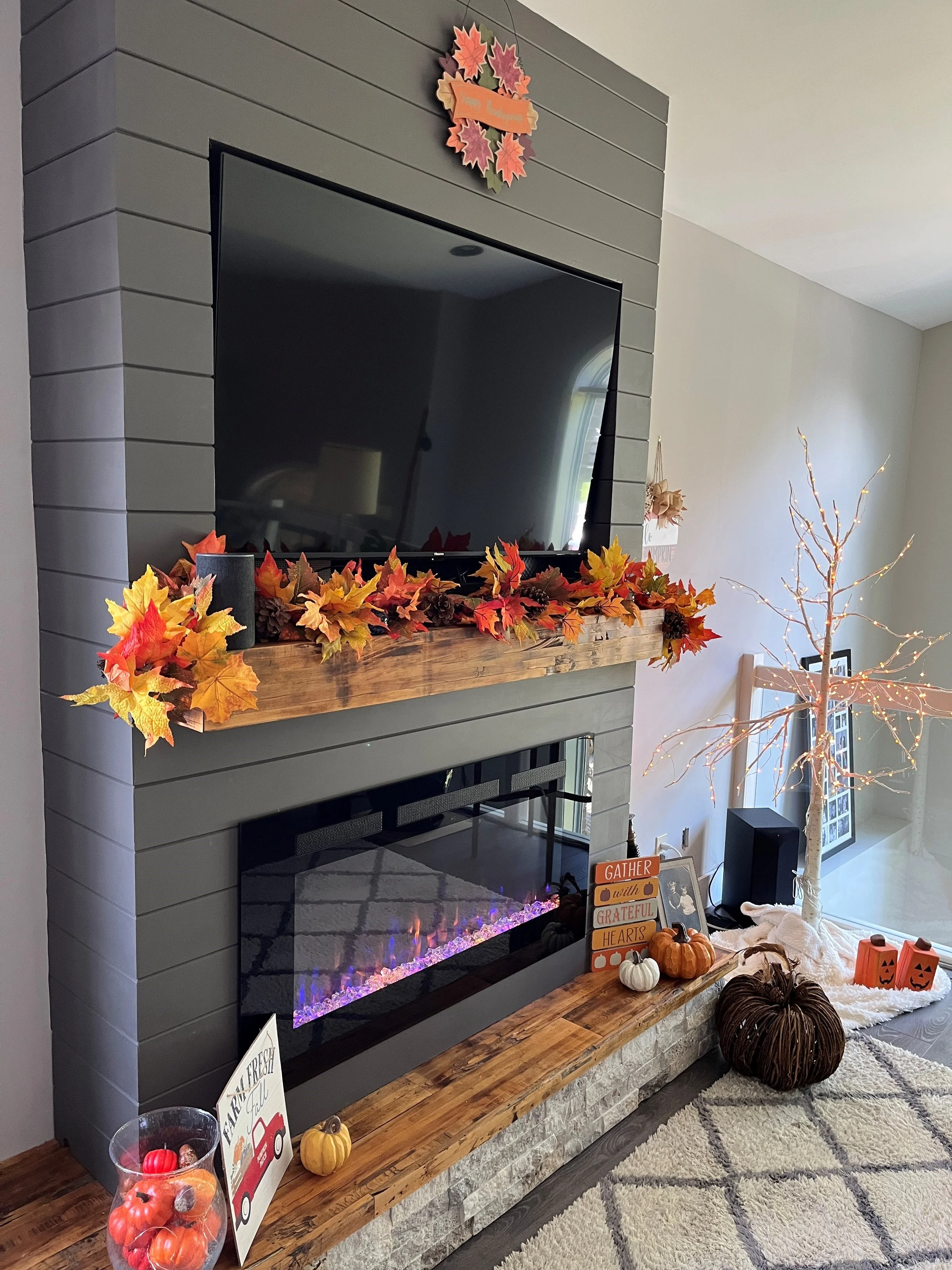 Decorative autumn-themed arrangement on a fireplace mantel with fall leaves, pumpkins, and candles in a cozy living room.