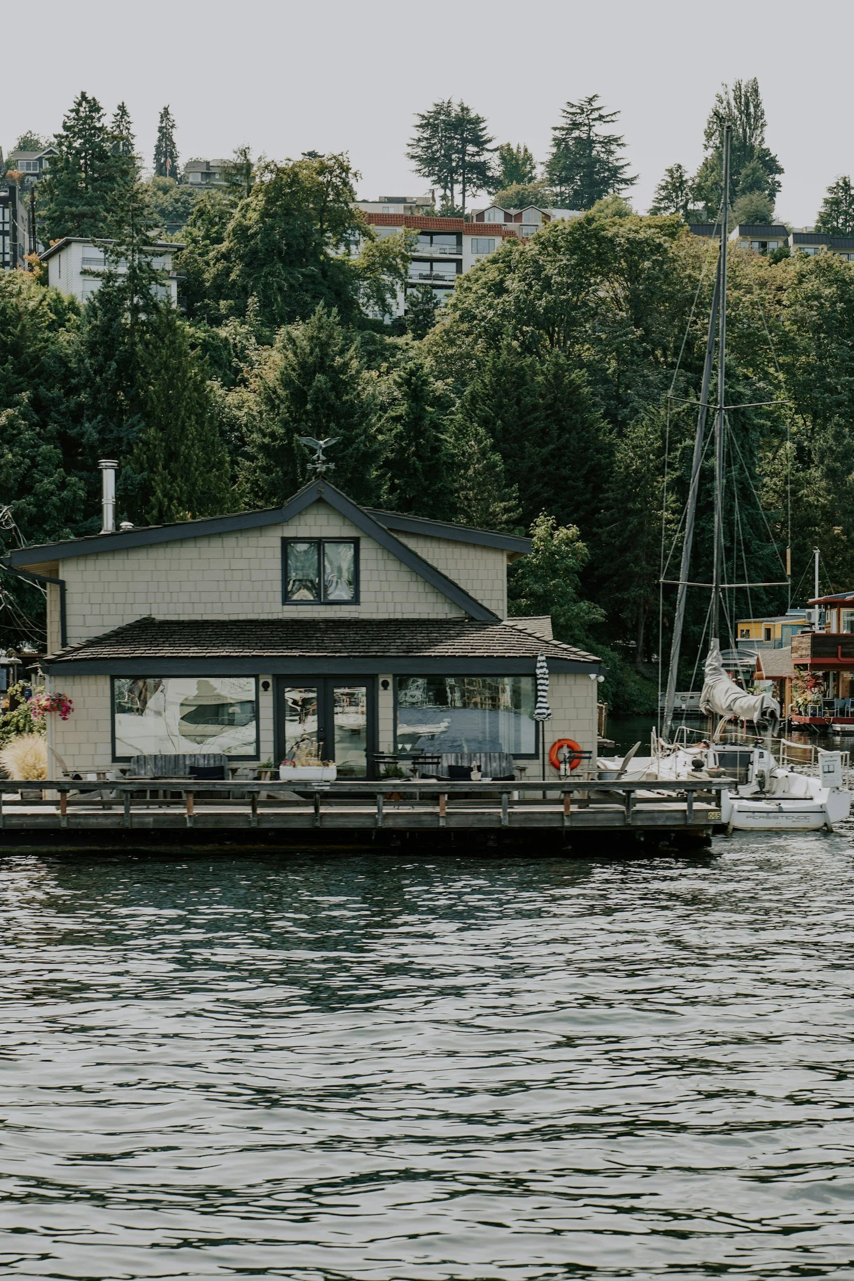 A house on a dock with a boat, surrounded by trees and water, with residential buildings on a hillside in the background.