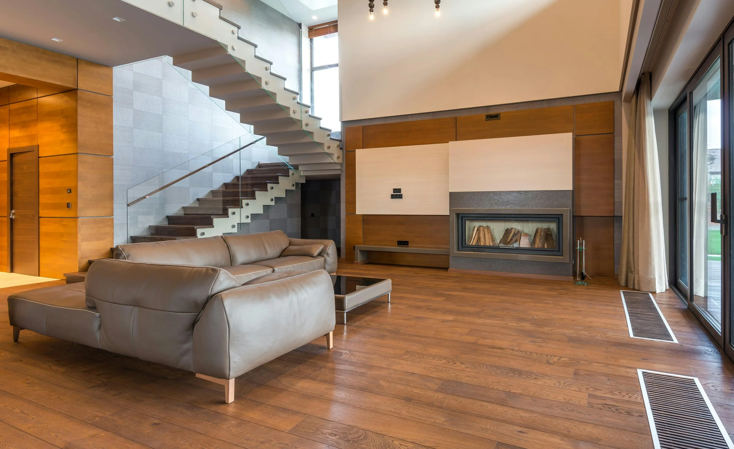 Modern living room with leather sofas, wooden flooring, a fireplace, large windows with curtains, and a staircase with glass railing leading upstairs.