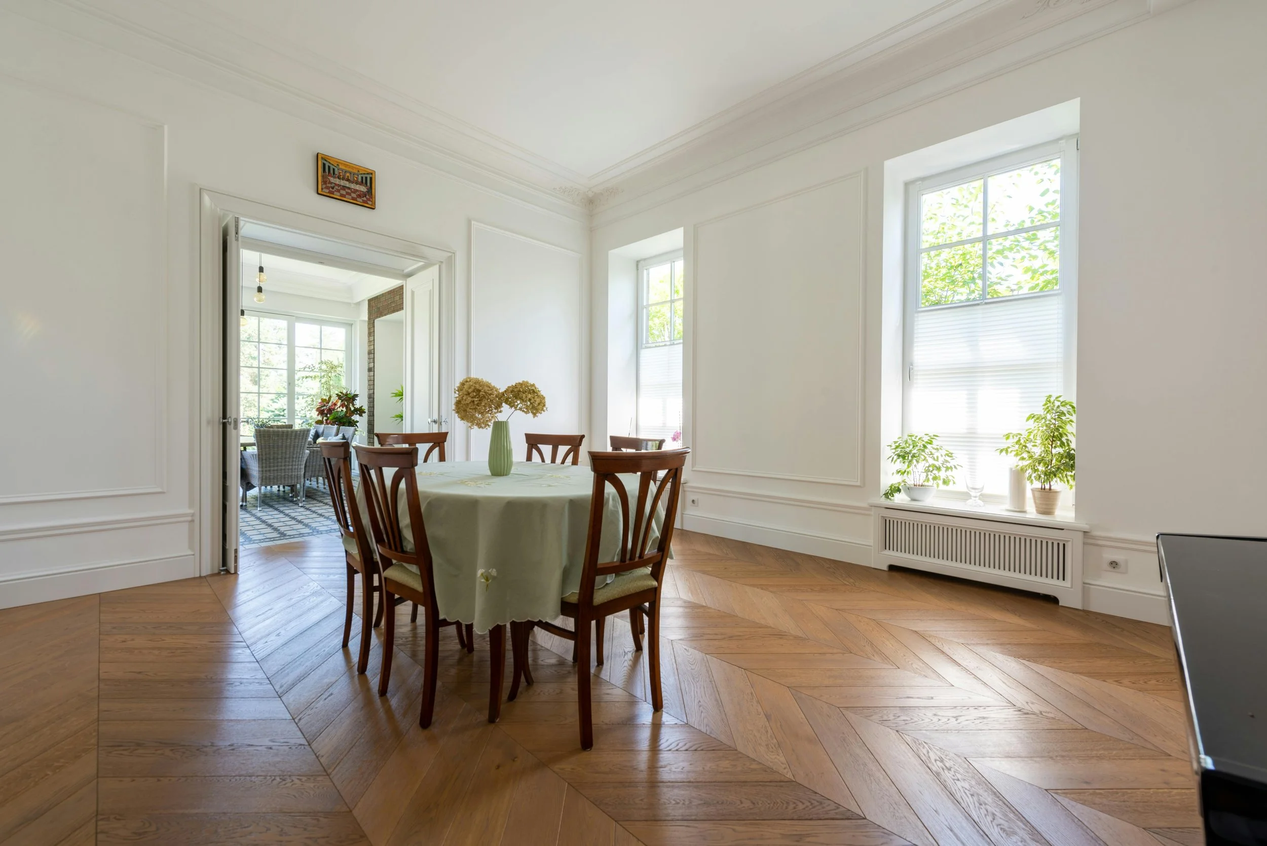A bright dining room with a round table and six wooden chairs, decorated with a vase of dried flowers. The room has large windows with white blinds, white walls with molding, wooden herringbone flooring, and potted plants on the windowsills.