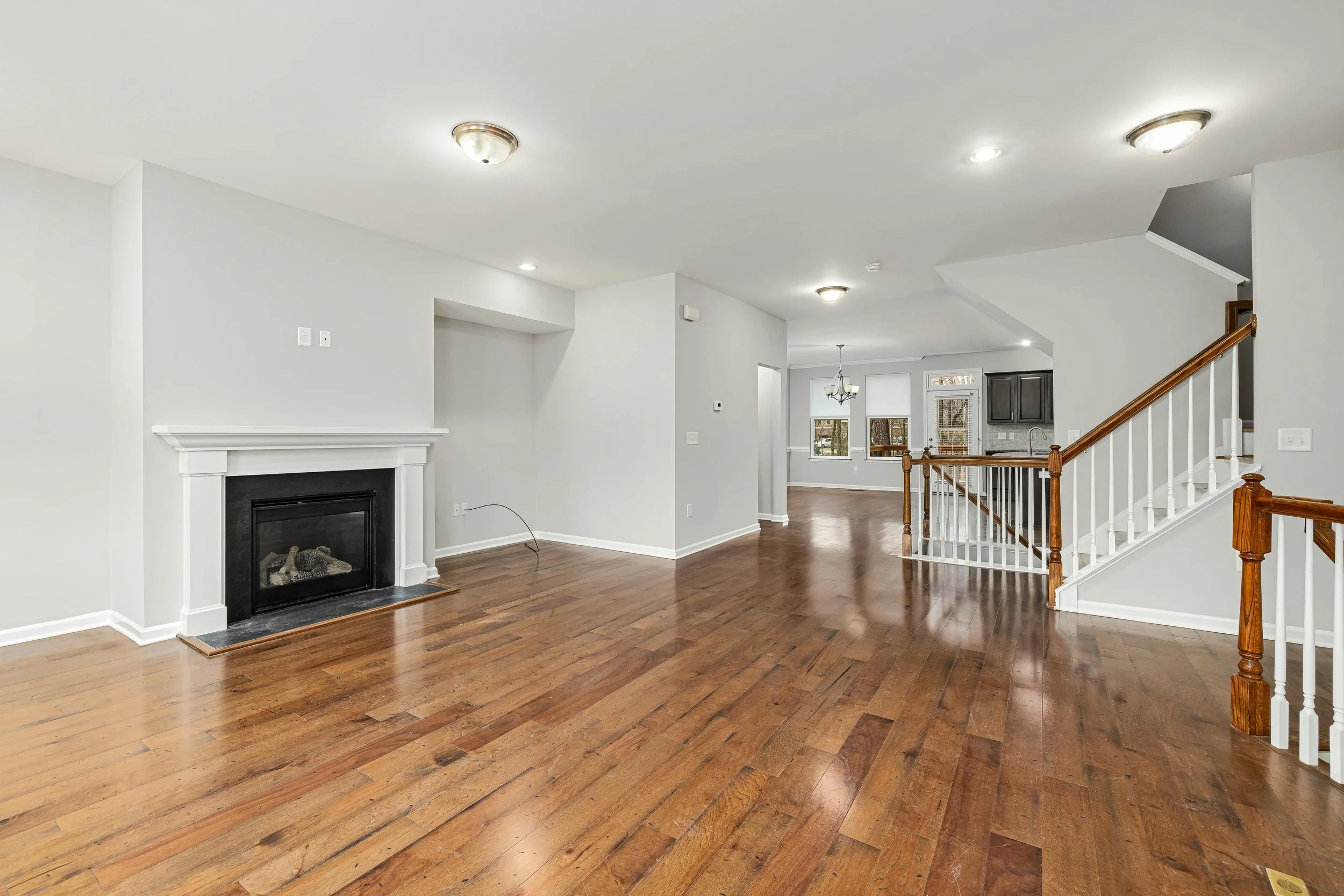 Empty living room with hardwood floors, white walls, a fireplace, and a staircase leading upstairs, with visible dining area and kitchen in the background.
