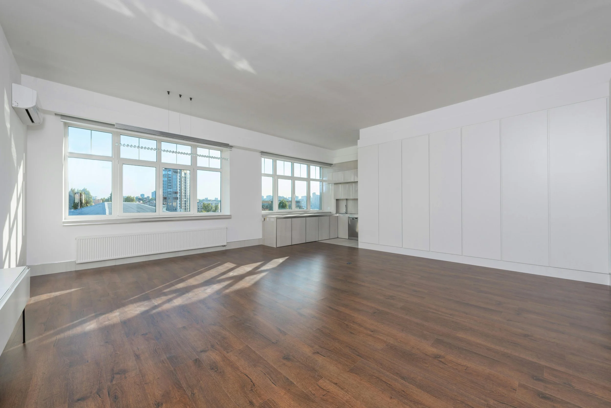 Empty living room with large windows, white walls, hardwood floors, and a small kitchen area in the corner.