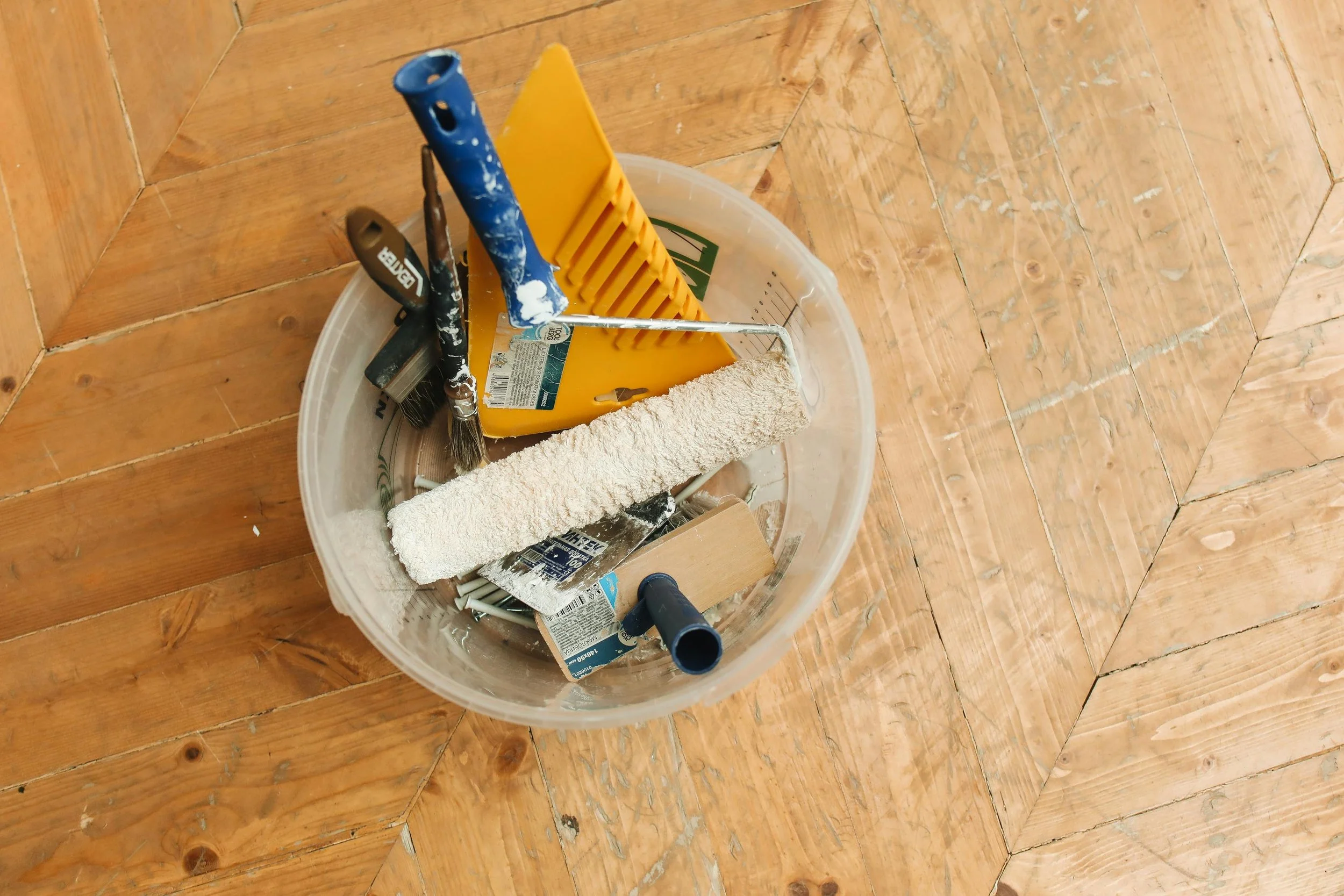 A plastic bucket containing painting tools including brushes, a paint roller, a paint scraper, a paint comb, and a paint tray, placed on a wooden floor.