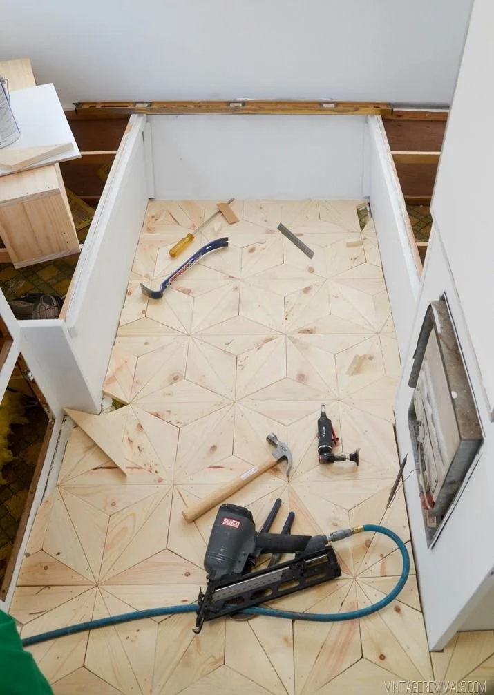 A kitchen under renovation with a partially installed tile floor, tools including a nail gun, hammer, screwdriver, and a square, and a utility knife on the floor, with some wooden boards and a sketch or blueprint on a side table.