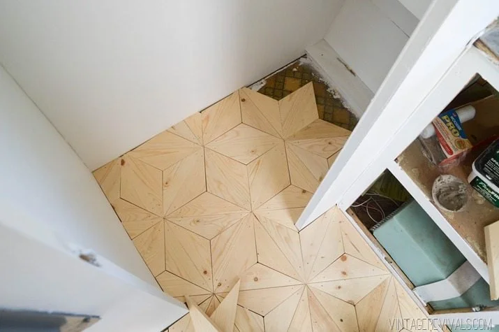 Room corner with newly installed geometric wood floor tiles and partially finished white walls, showing construction or renovation in progress.