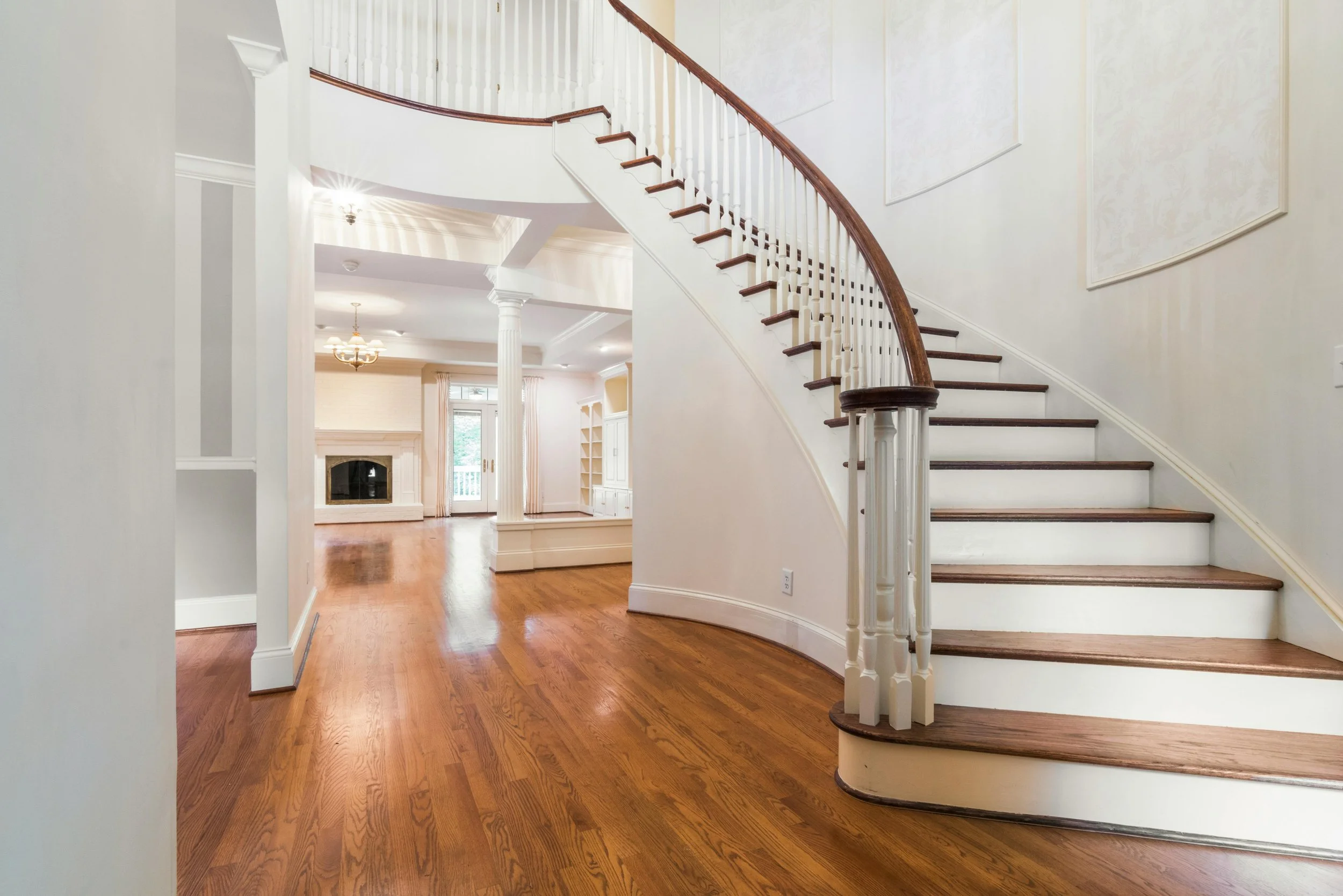 Interior view of a spacious house with hardwood floors, a curved staircase with white risers and wooden treads, and a living room in the background featuring a fireplace, large windows, and a chandelier.