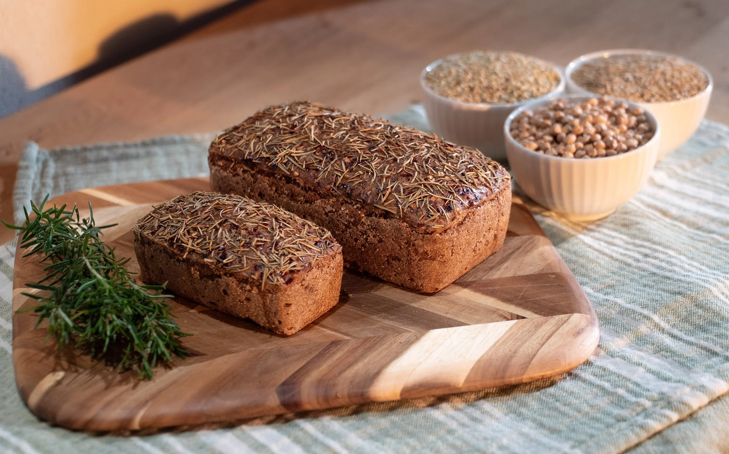Two loaves of bread topped with rosemary on a wooden cutting board, with sprigs of rosemary nearby. Three small bowls filled with various seeds or grains in white bowls in the background.