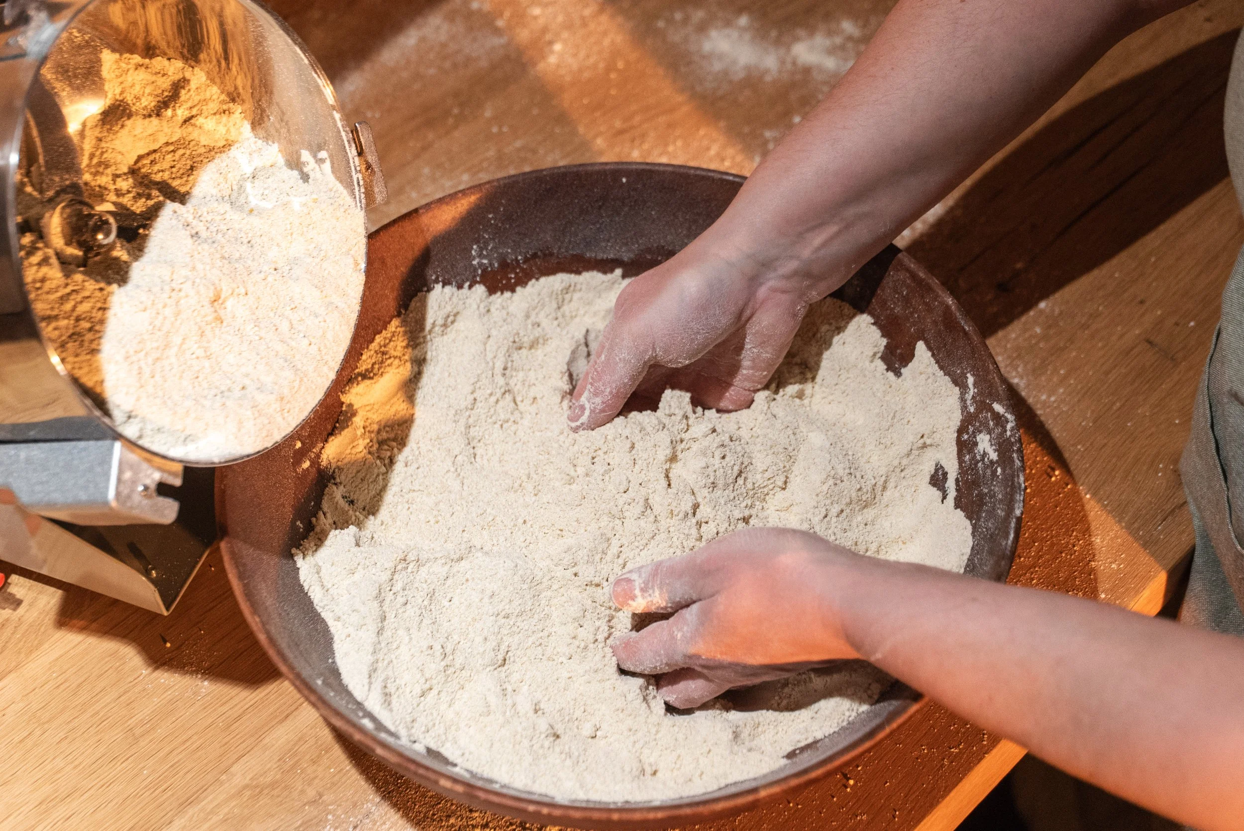 Person kneading dough in a bowl with flour, nearby a metal container with more flour on a wooden surface.