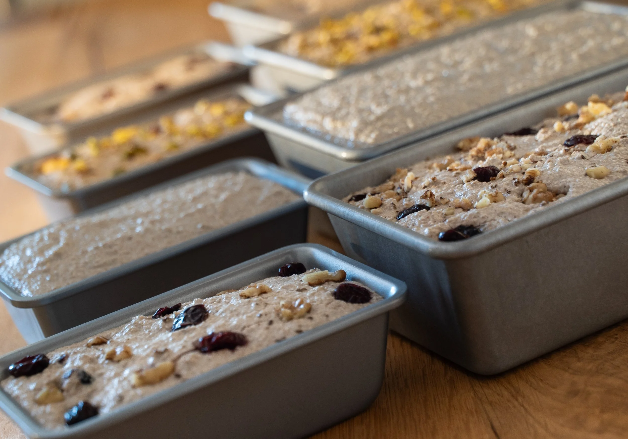 Multiple loaf pans filled with unbaked artisanal sourdough bread made with organic gluten-free ingredients, some topped with dried cranberries and chopped nuts, set on a wooden surface.
