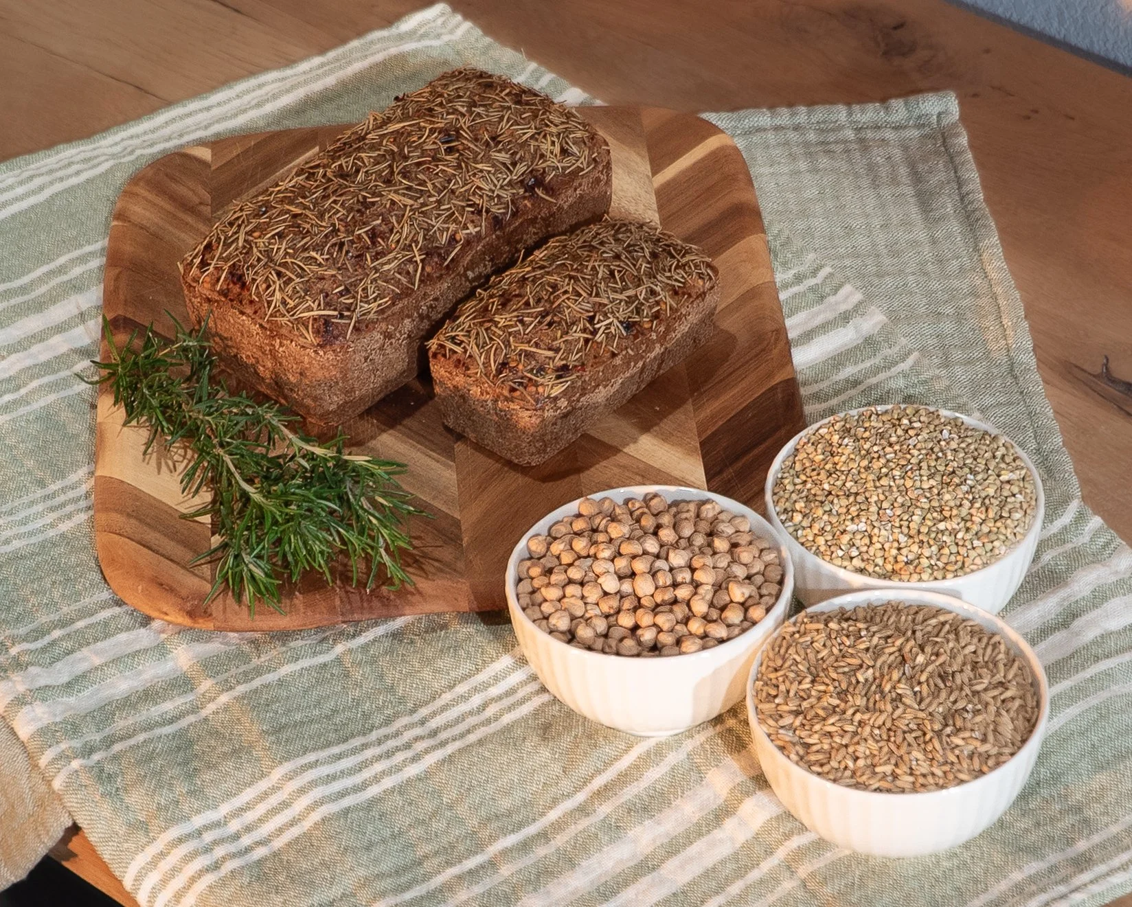 Two loaves of bread topped with rosemary on a wooden cutting board, three bowls of different grains, and a sprig of rosemary on a striped tablecloth.