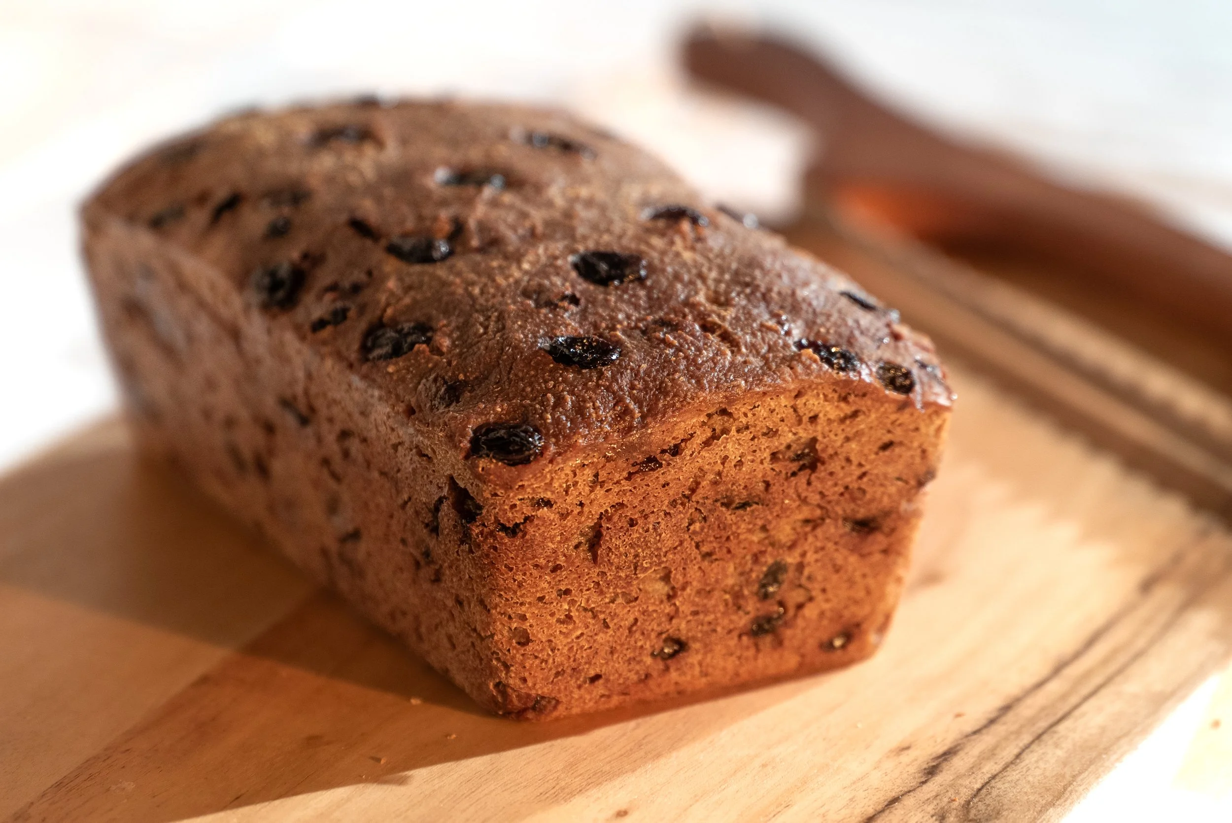 Close-up of a freshly baked Raisin Cinnamon Sourdough Bread Loaf with organic gluten-free ingredients on a wooden cutting board, with a bread knife in the background.