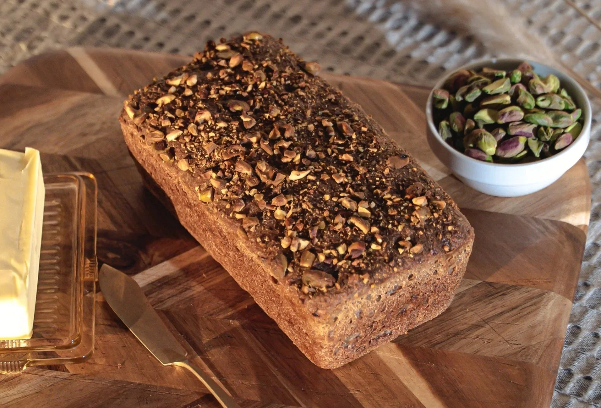 A Loaf of Pistachio Sourdough Bread with organic gluten-free ingredients topped with chopped pistachios on a wooden cutting board, with a small bowl of pistachios nearby.