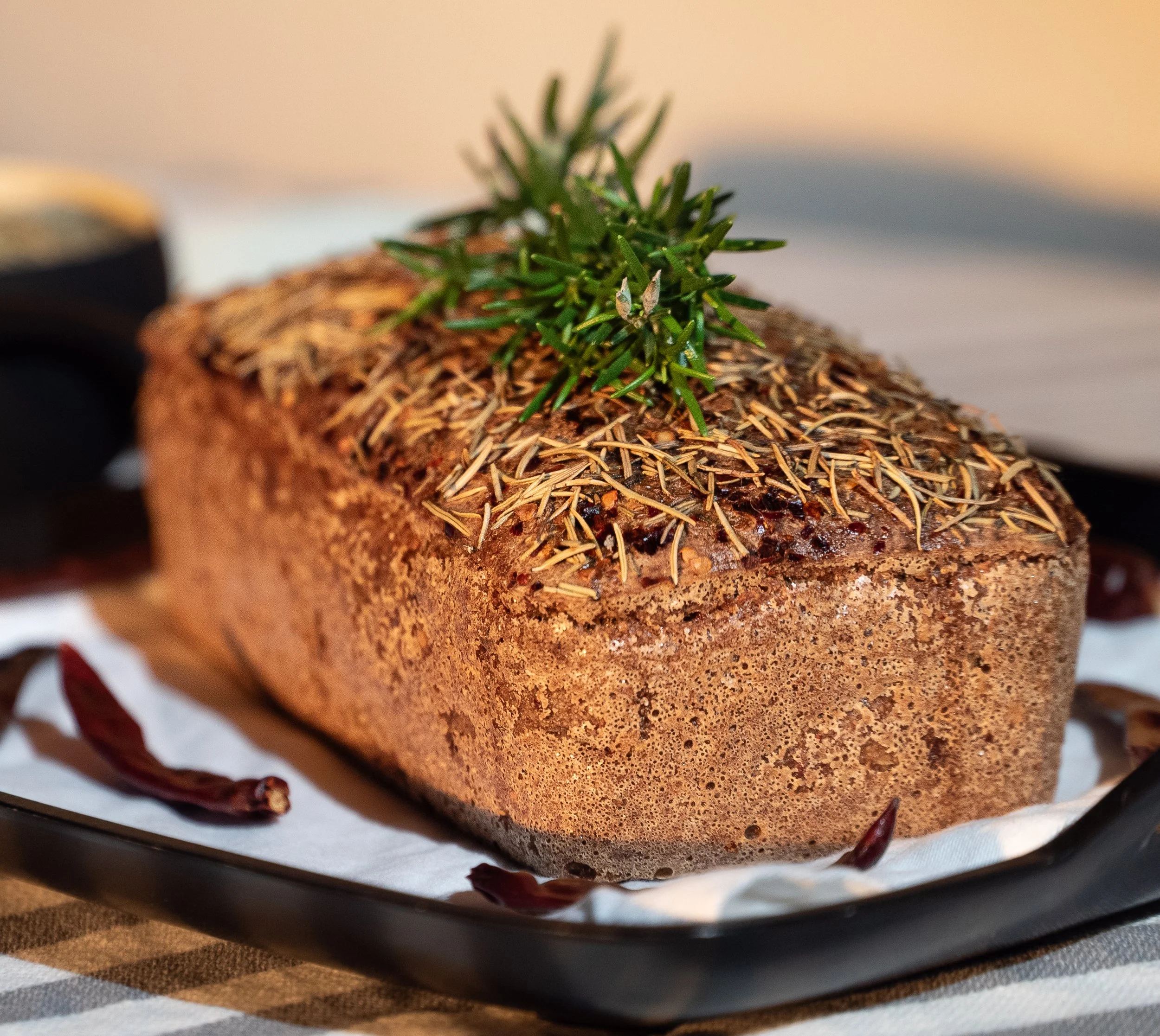 Freshly baked bread loaf garnished with rosemary and dried herbs, on a baking tray with dried chili peppers around, on a striped tablecloth.