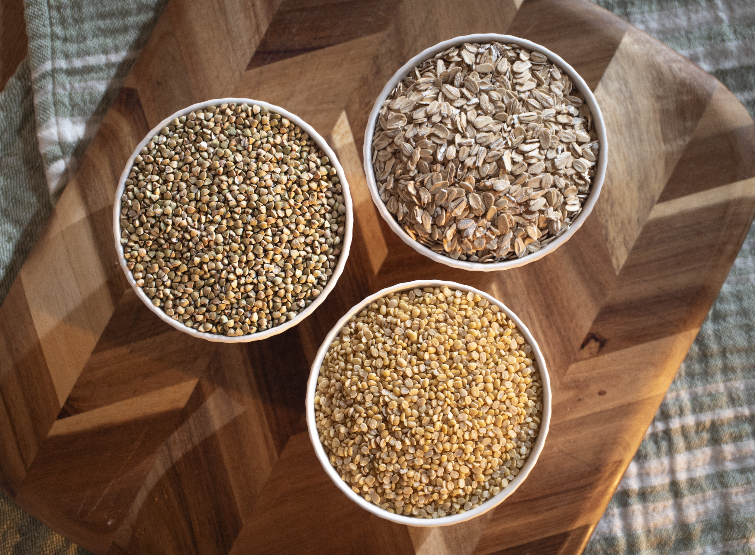 Three bowls filled with different types of organic buckwheat groats, organic rolled oats, organic mung dal beans on a wooden cutting board.
