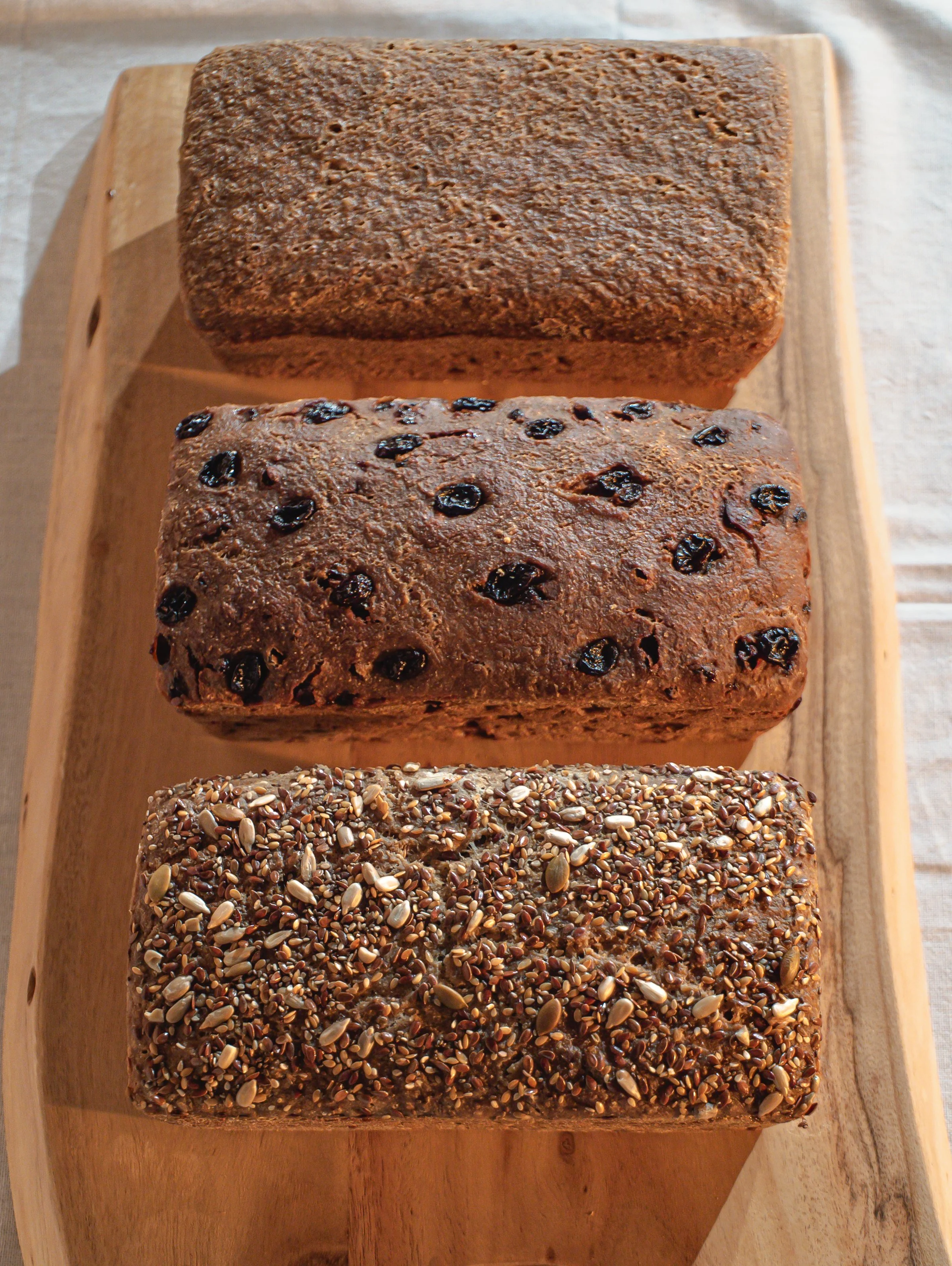 Three loaves of sourdough bread made with organic gluten-free ingredients on a wooden serving board, each with different toppings: Classic, Cinnamon Raisin, and Six Seeds mixture.