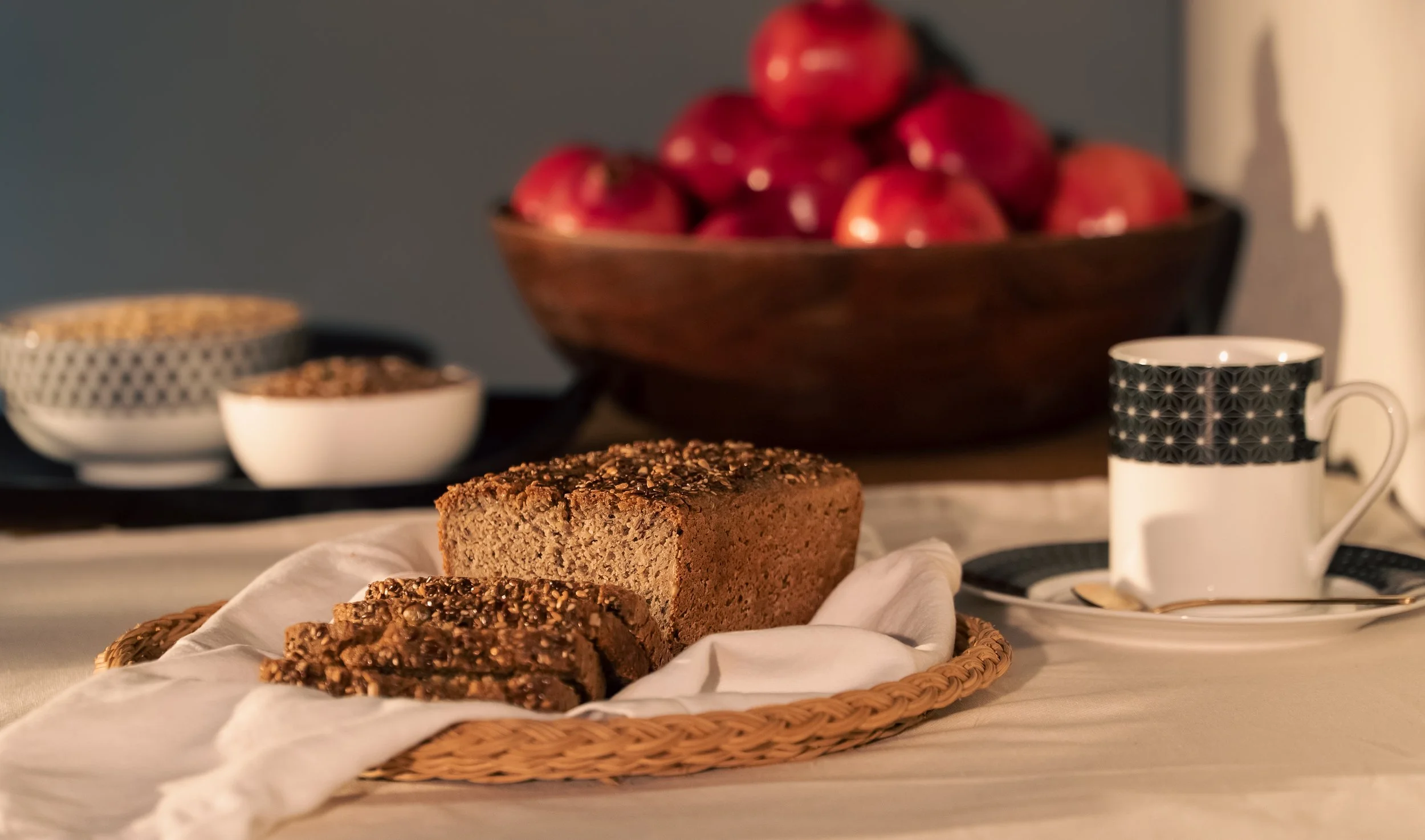 A loaf of sourdough bread made with organic gluten-free ingredients on a white cloth with slices cut, on a woven basket. In the background, a large wooden bowl filled with red pomegranates, white bowls with toppings, a cup and saucer with a spoon.