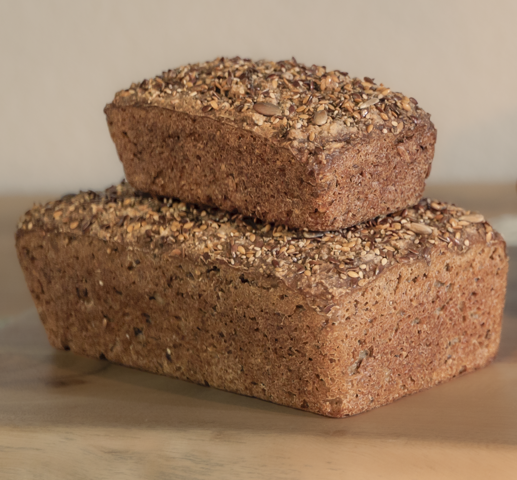 Two loaves of Six Seeds Loaf and Mini Loaf bread made with organic gluten-free ingredients, stacked, topped with seeds, on a wooden surface.