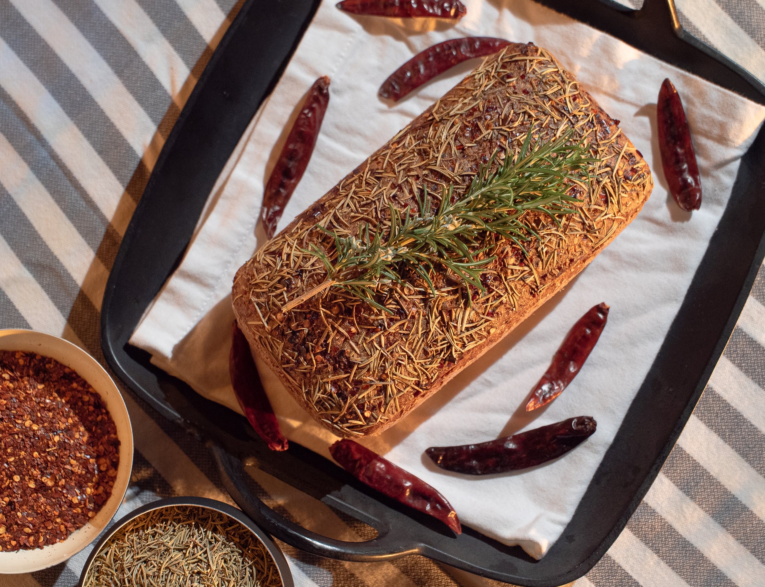 Herb crusted roast beef garnished with a sprig of rosemary on a baking sheet, surrounded by dried red chili peppers and bowls of spices.