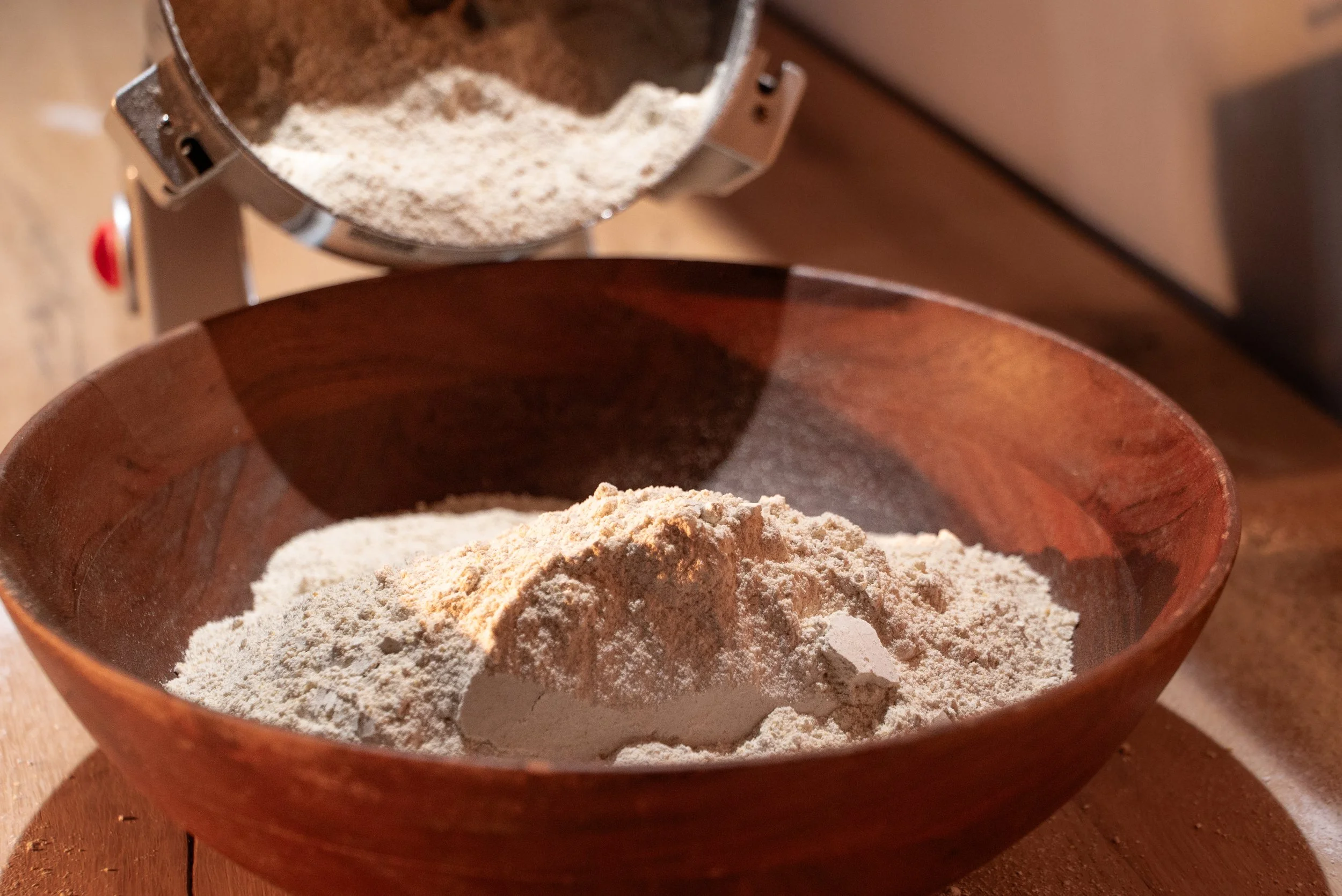 Close-up of a wooden bowl filled with flour on a wooden surface, with a metal container pouring more flour into it.