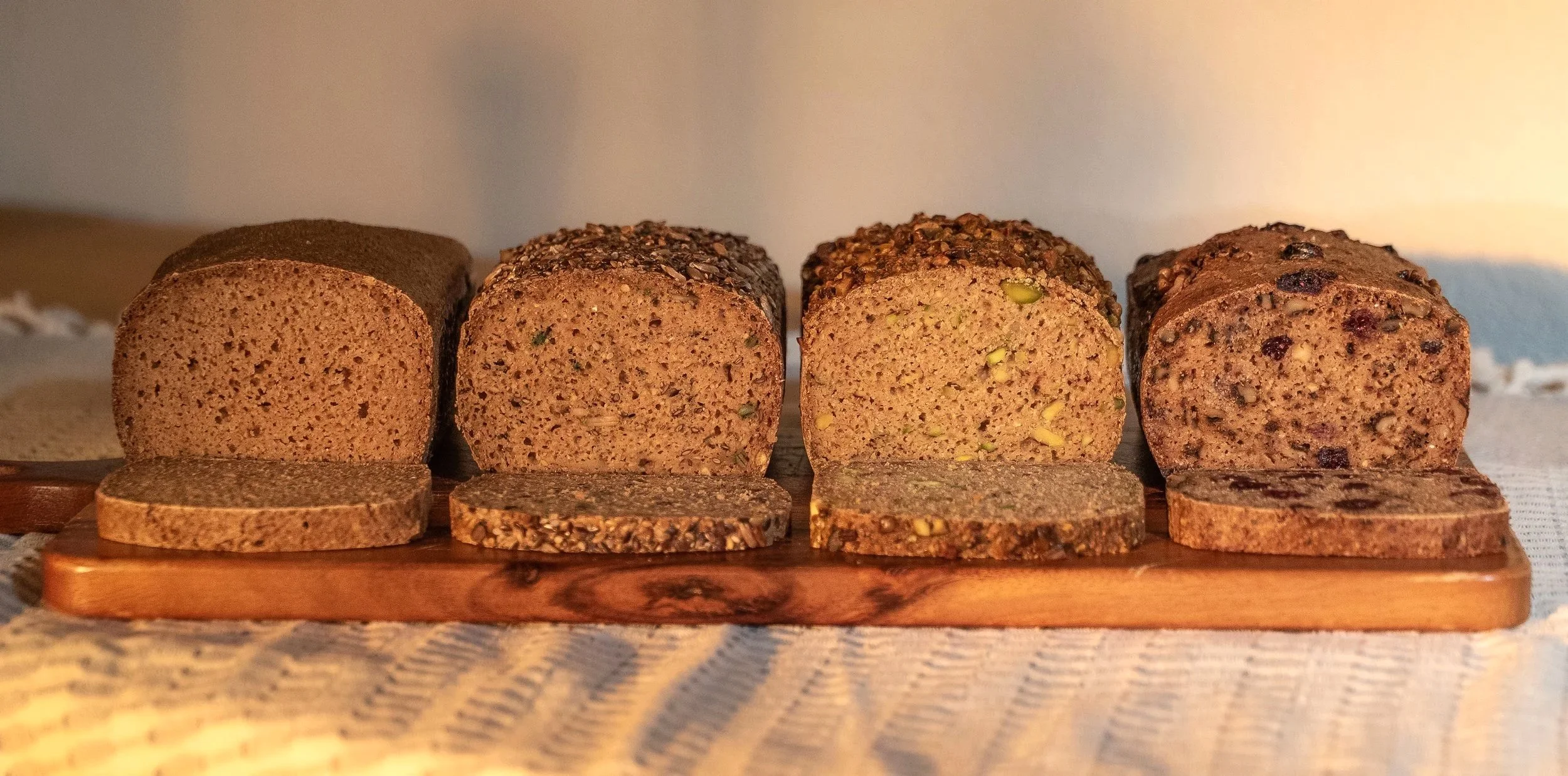 Four loaves of multigrain bread on a wooden board, with slices cut and placed in front, showcasing different textures and seed toppings.