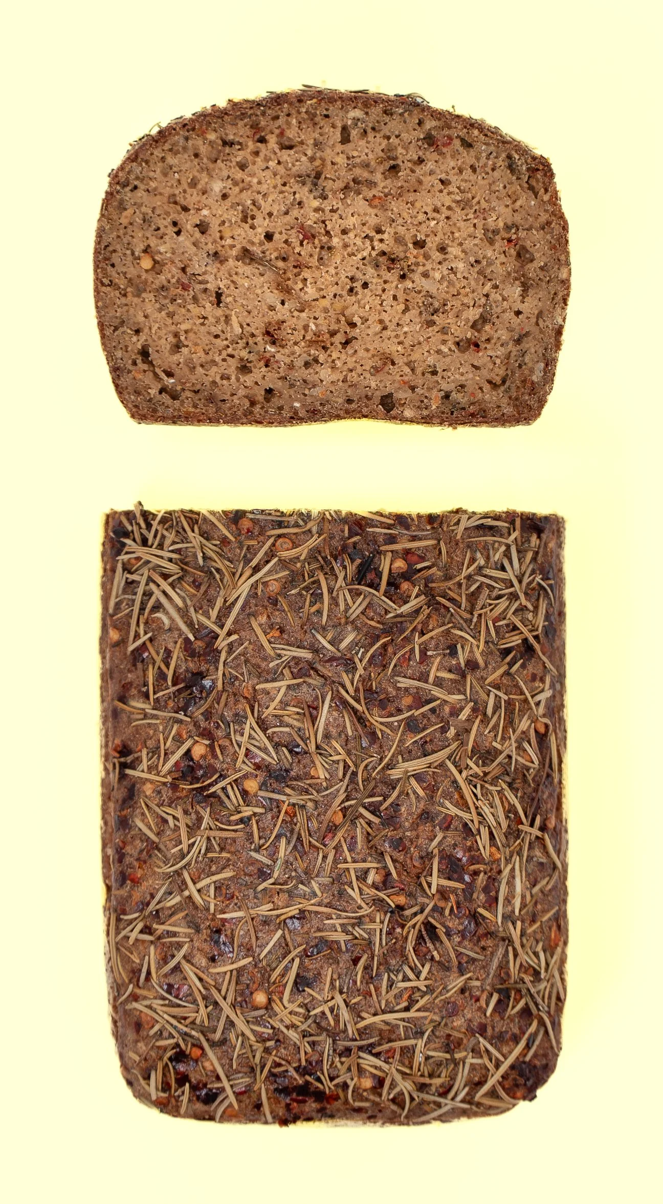 Two slices of rye bread, one plain and one topped with rosemary and spices, on a light yellow background.