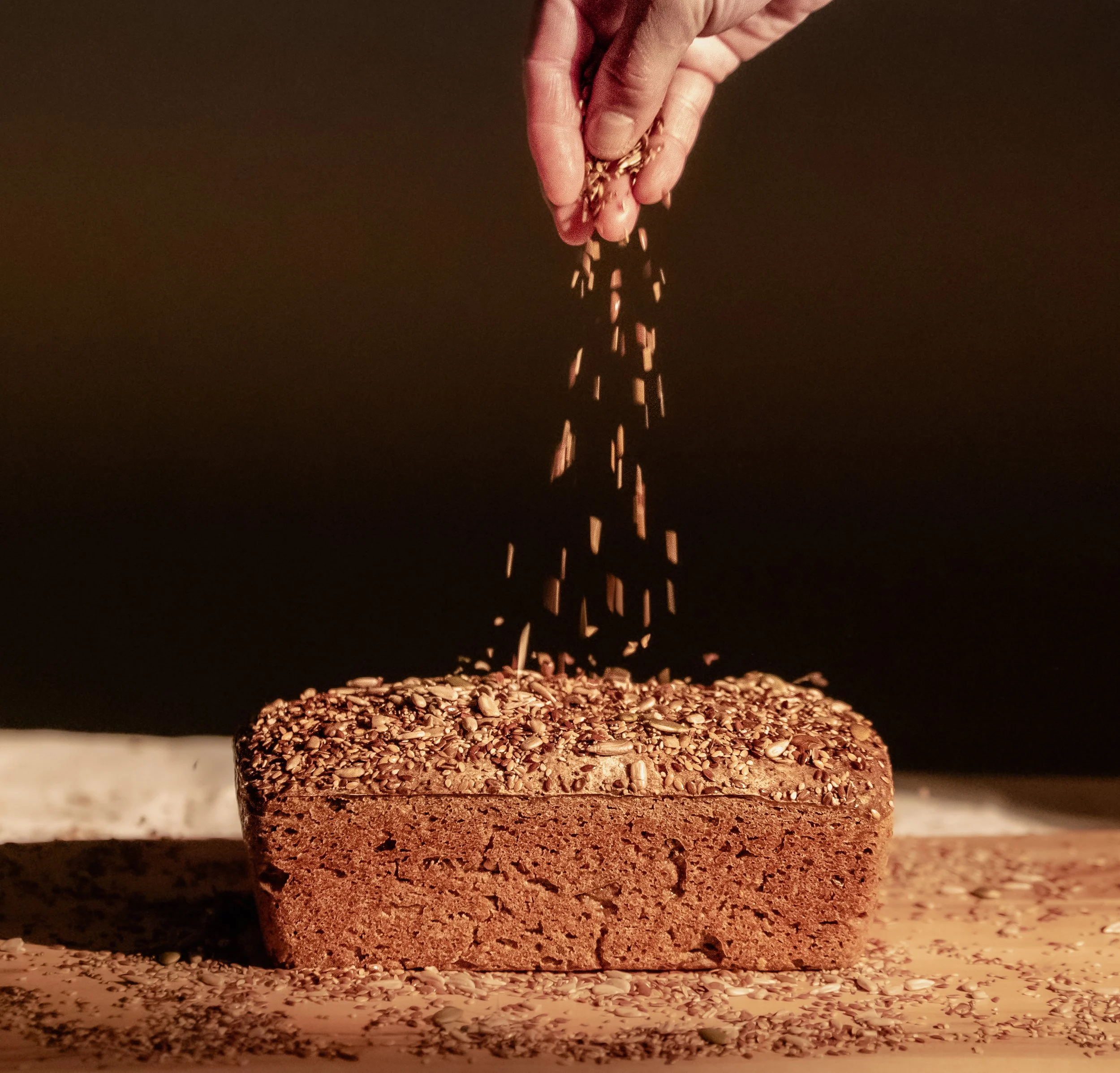 A hand sprinkling seeds over a freshly baked loaf of Six Seeds Sourdough bread made with organic gluten-free ingredients, topped with seeds, on a wooden surface with grains scattered around, against a dark background.
