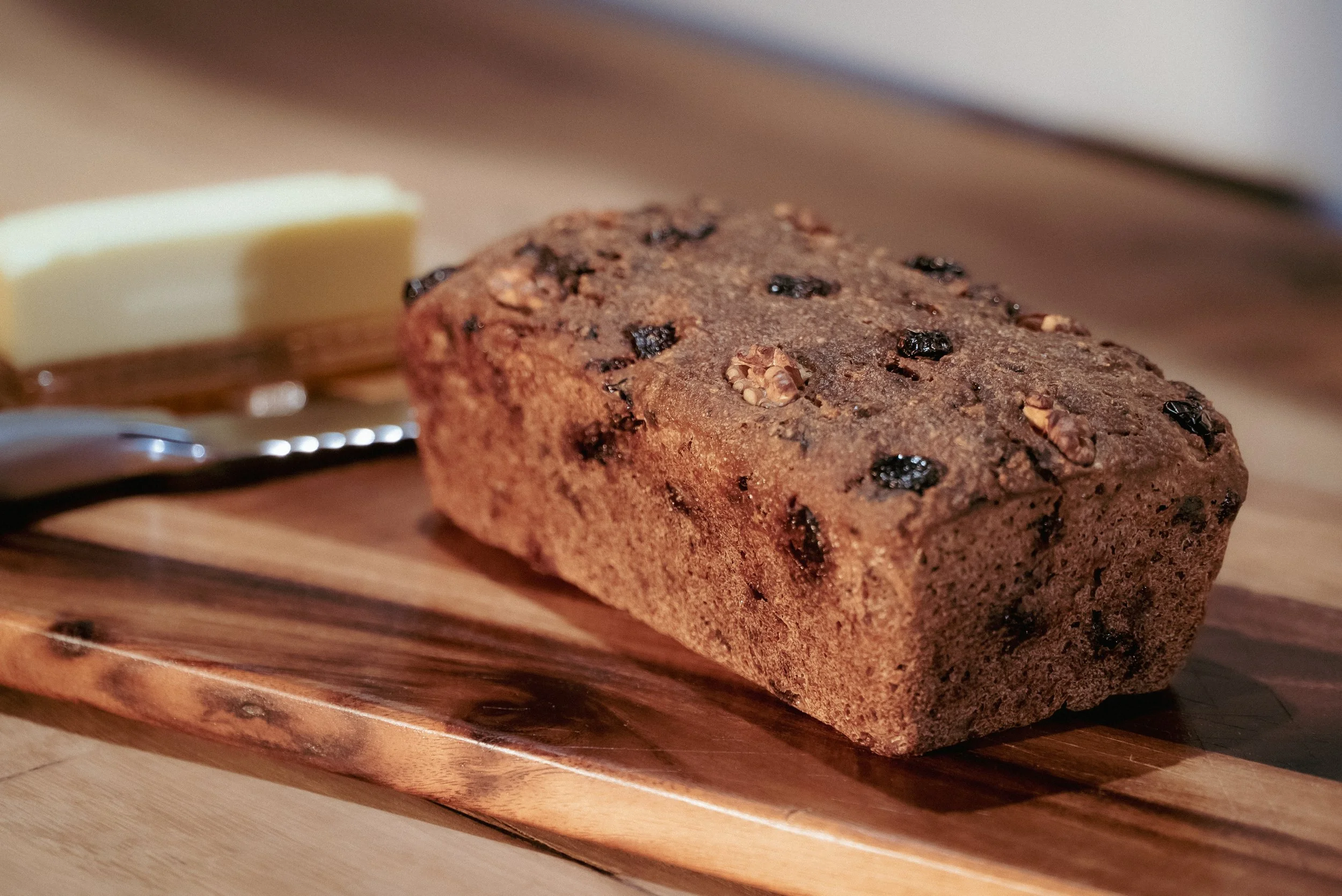 A Cranberry Walnut Sourdough Loaf Bread with organic gluten-free ingredients on a wooden cutting board with a pats of butter, a butter knife, and a butter spreader in the background.