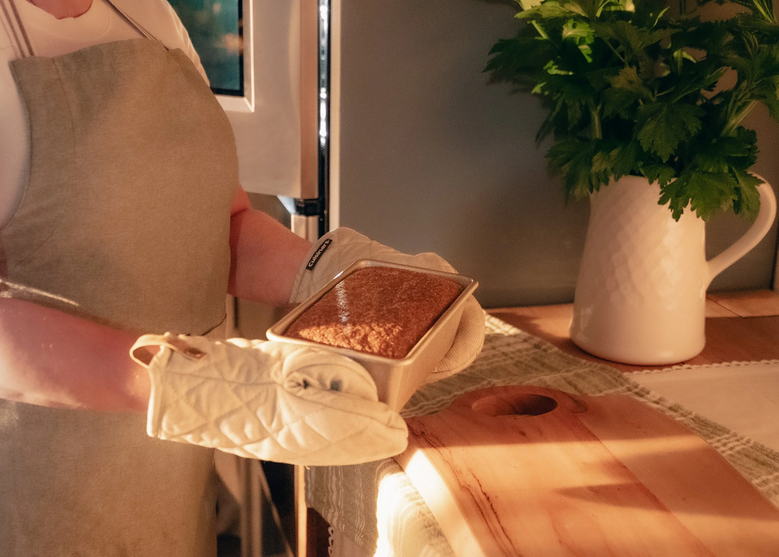 A person wearing oven mitts holding a freshly baked artisan bread made with organic gluten-free ancient grains and legumes in a pan.  In the background, there is a large white pitcher with green foliage and a wooden table with a woven placemat.