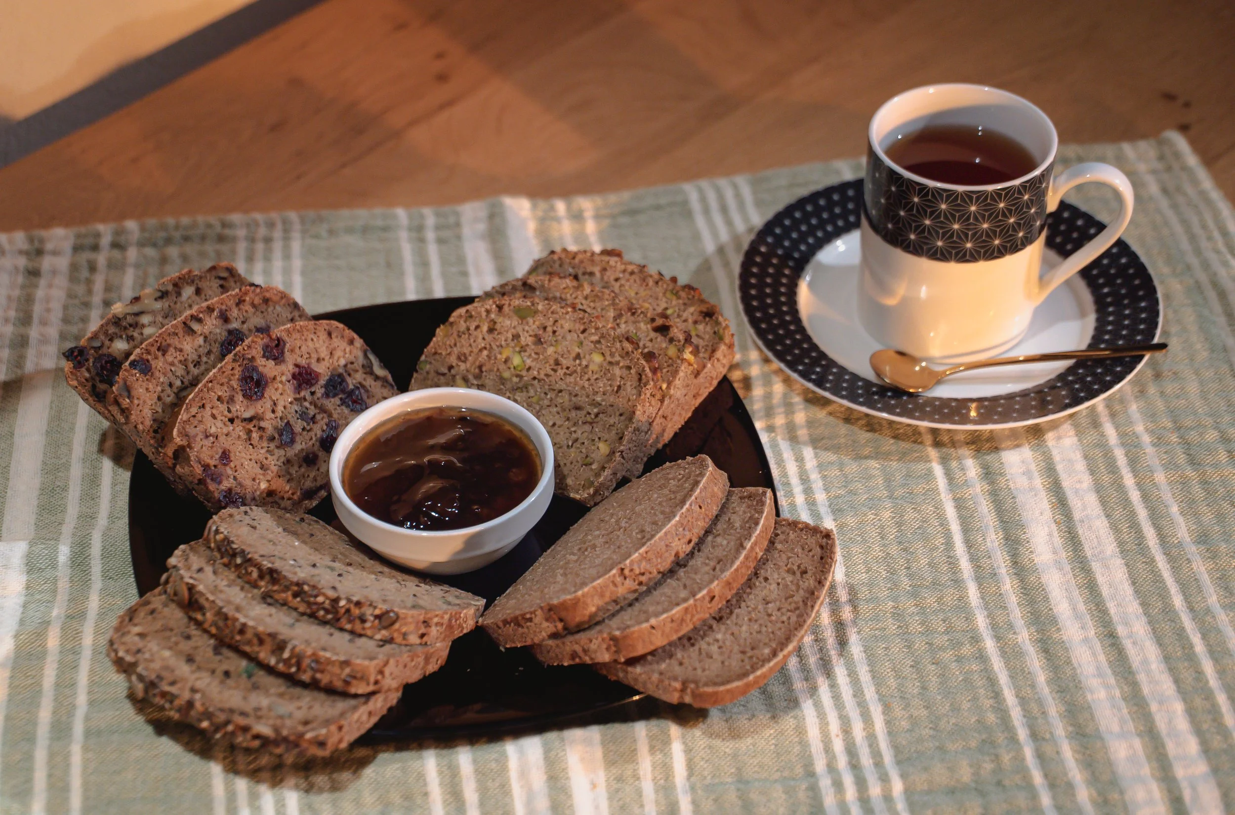 Assorted slices of multigrain bread on a black plate with a small bowl of jam, served alongside a cup of tea on a white cup with black rim and a small golden spoon, all on a checkered beige placemat.