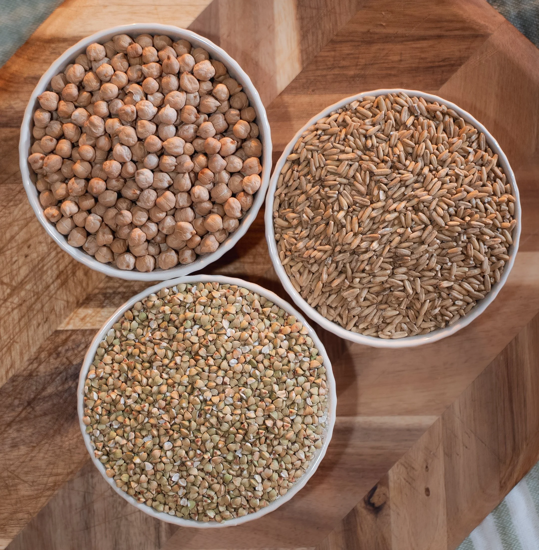 Three white bowls filled with different types of grains placed on a wooden cutting board.