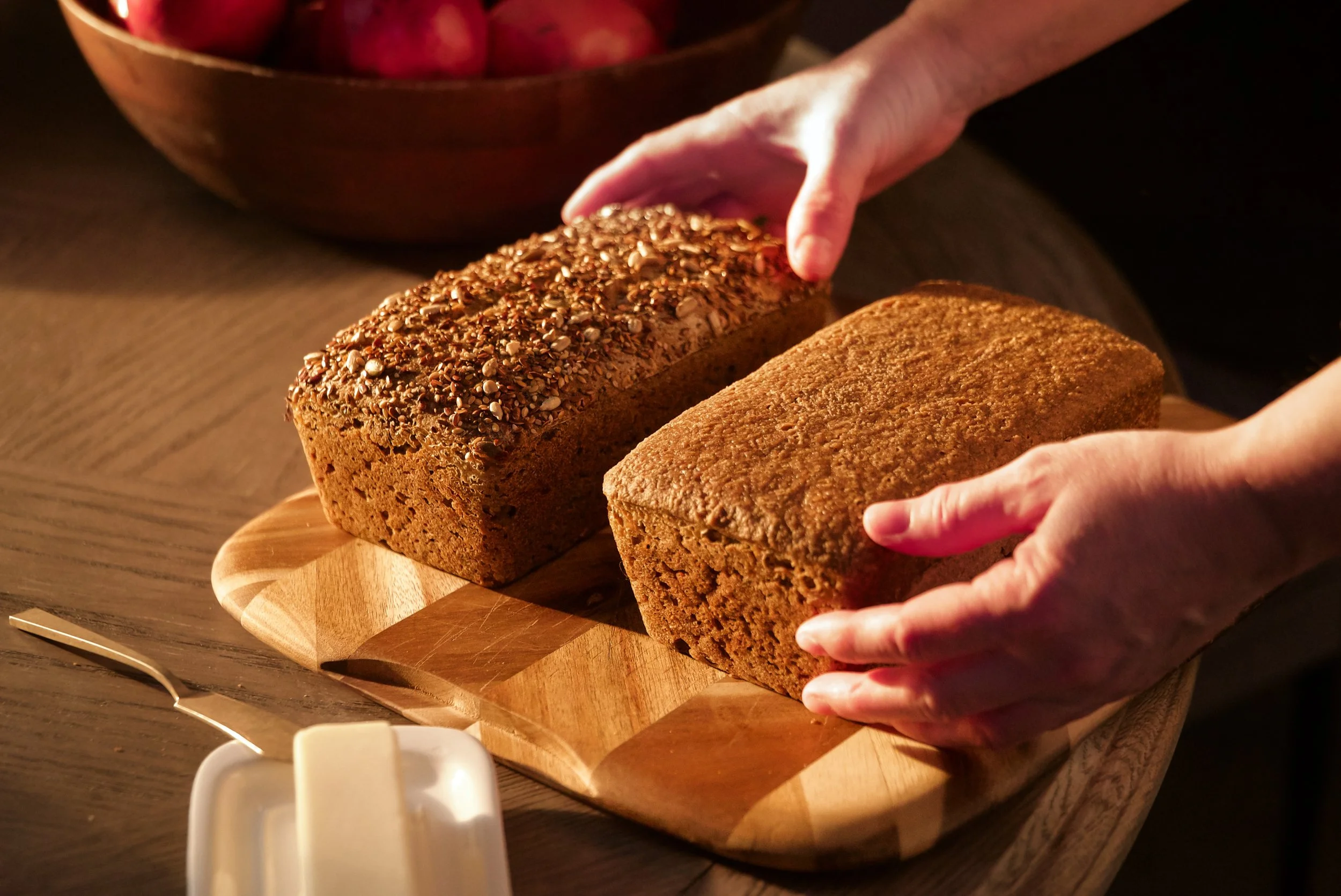 Two whole loaves of bread, one topped with seeds and grains, on a wooden cutting board with hands holding each loaf, in a cozy kitchen setting.