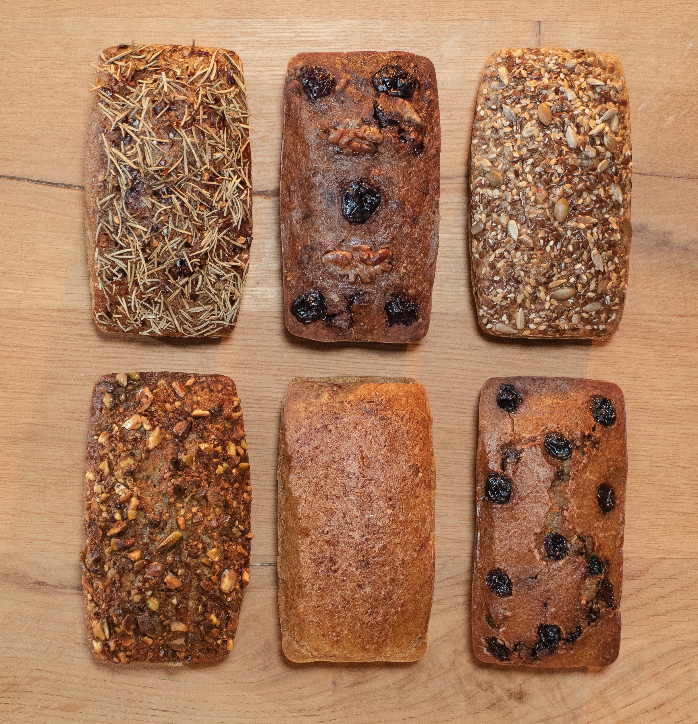 Six different rectangular loaves of bread decorated with various toppings, arranged on a wooden surface.
