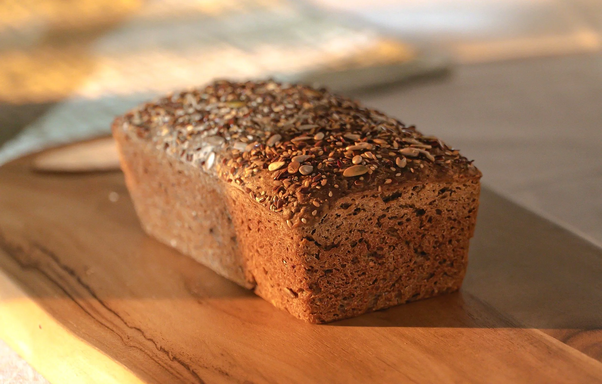 An artisan sourdough bread loaf of whole organic ancient grain and legumes bread topped with mixed seeds, resting on a wooden cutting board with a blurred background.