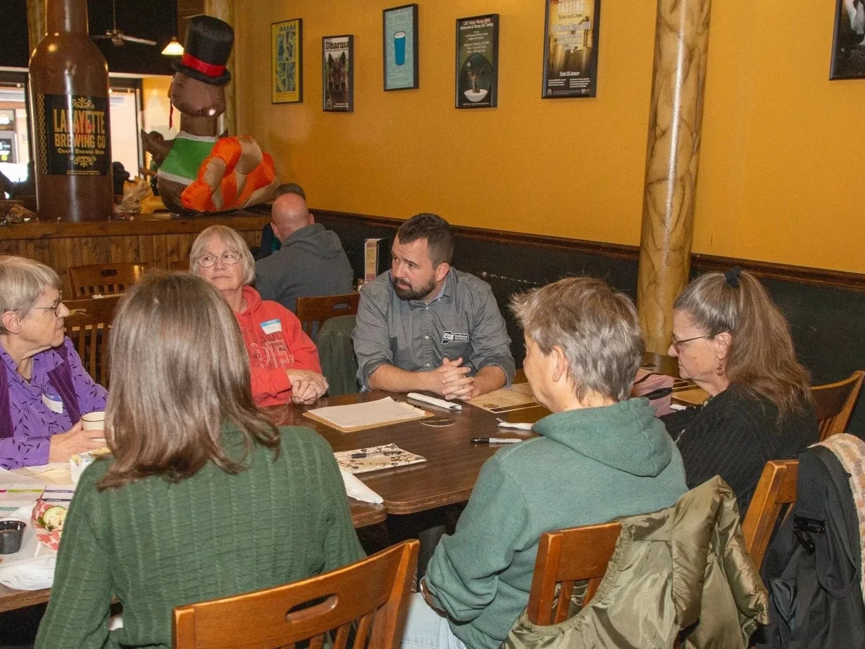 A group of people sitting around a table in a restaurant or café, engaged in conversation. The background shows a yellow wall with framed posters and décor, including a large inflatable stuffed animal wearing a top hat and a bow tie, along with a beer tap labeled La Porte Brewing Co.