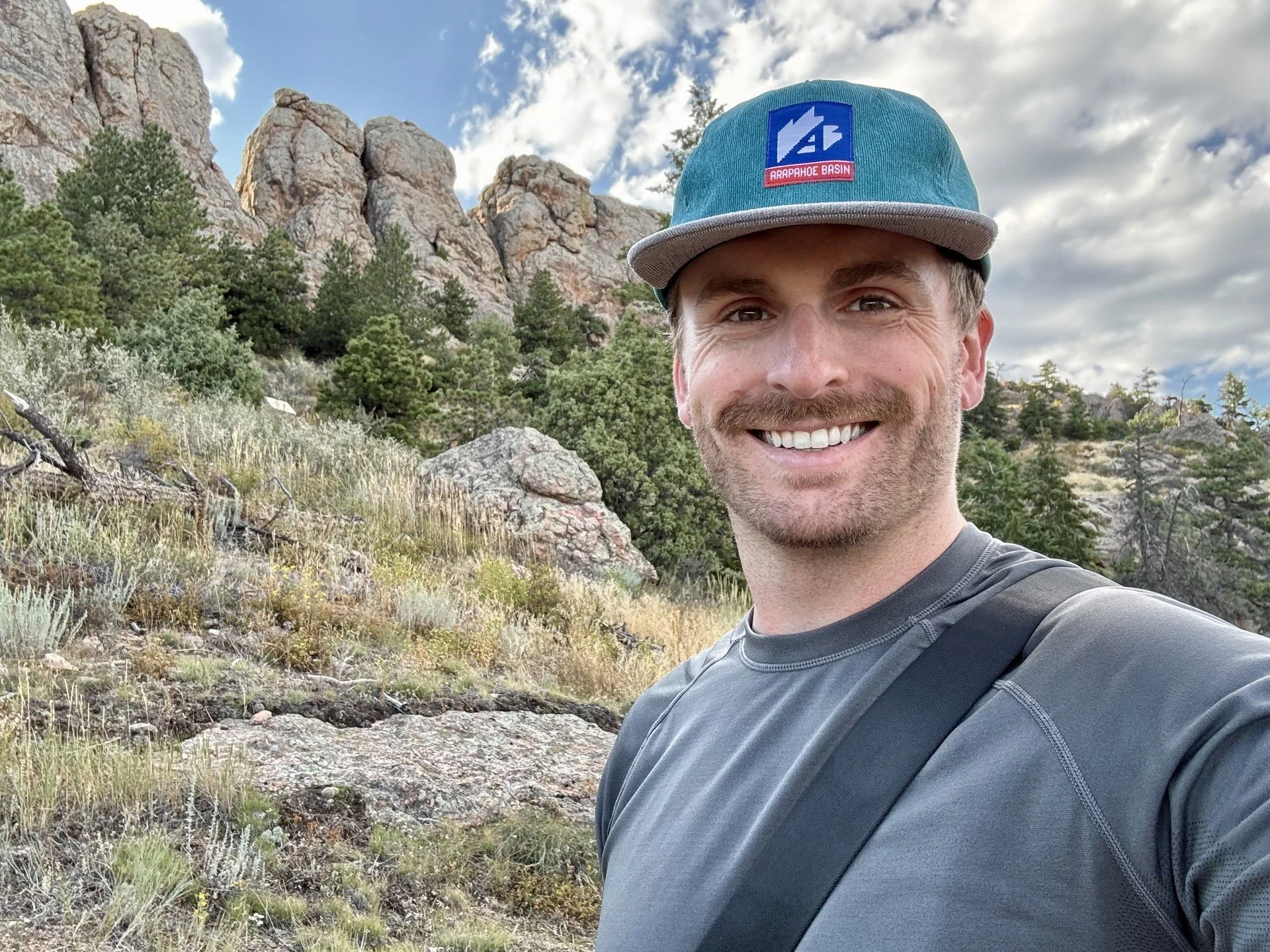 A man smiling outdoors in a mountainous area with trees and rocks, wearing a blue cap with a logo and a gray shirt.