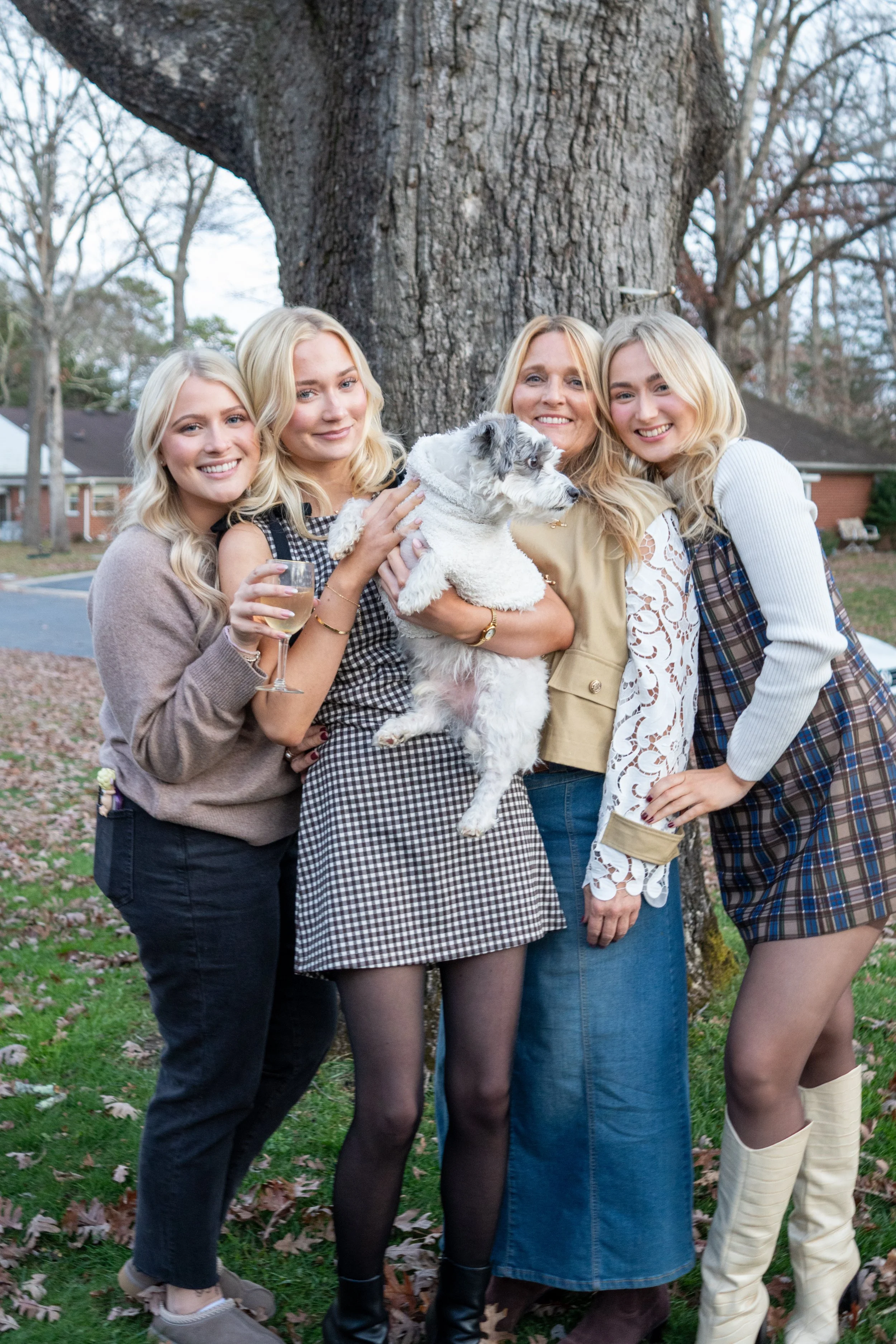 Four women and a dog standing outdoors in front of a large tree, smiling and posing for the camera.