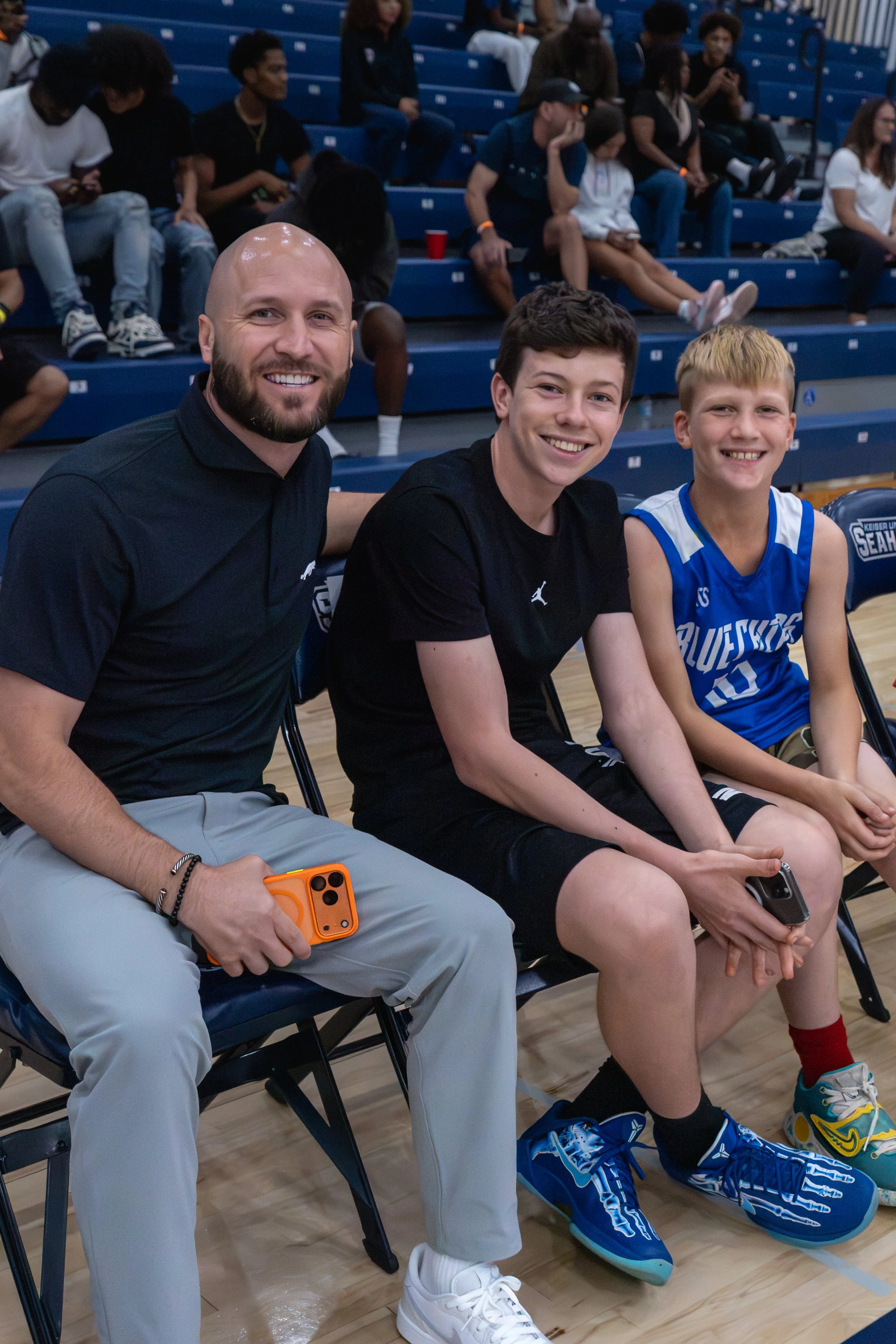 Three males sitting on chairs at a basketball game, smiling at the camera. The man on the left is bald with a beard, wearing a black shirt and gray pants, holding an orange phone. The young man in the middle has dark hair, wearing a black T-shirt and shorts, holding a phone. The young boy on the right has blond hair, wearing a blue basketball jersey and shorts, holding a phone. In the background, spectators are sitting on blue bleachers.