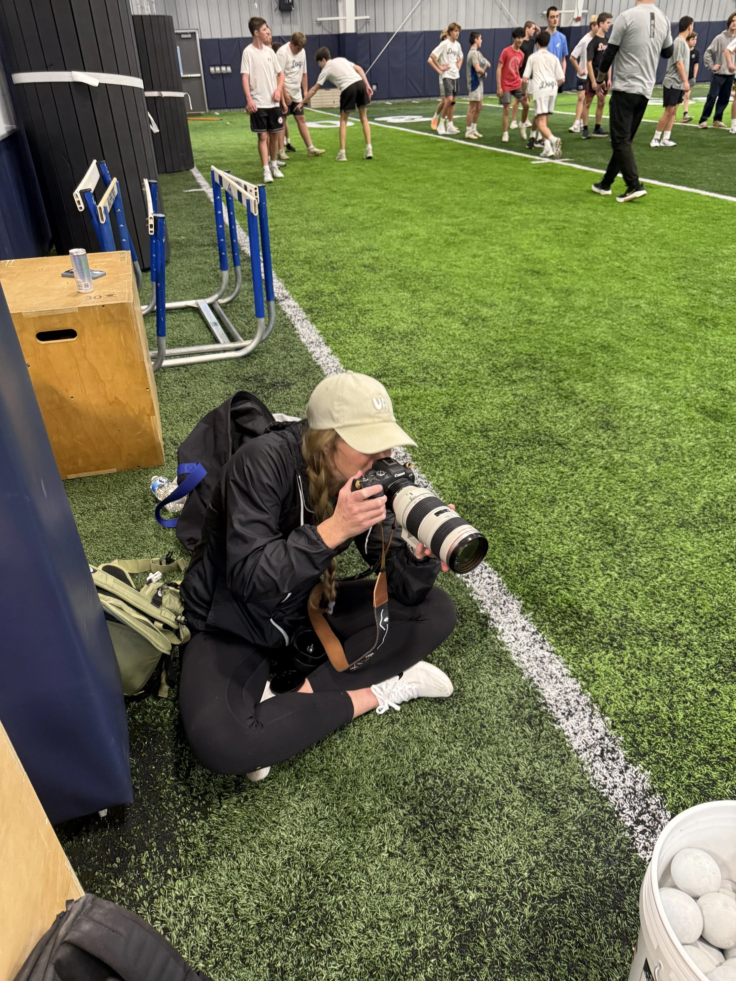 Coach Sam Mayer sitting on the floor taking a photograph with a professional camera at an indoor sports facility. There are children and adults in the background, some standing and some walking on the artificial turf field.