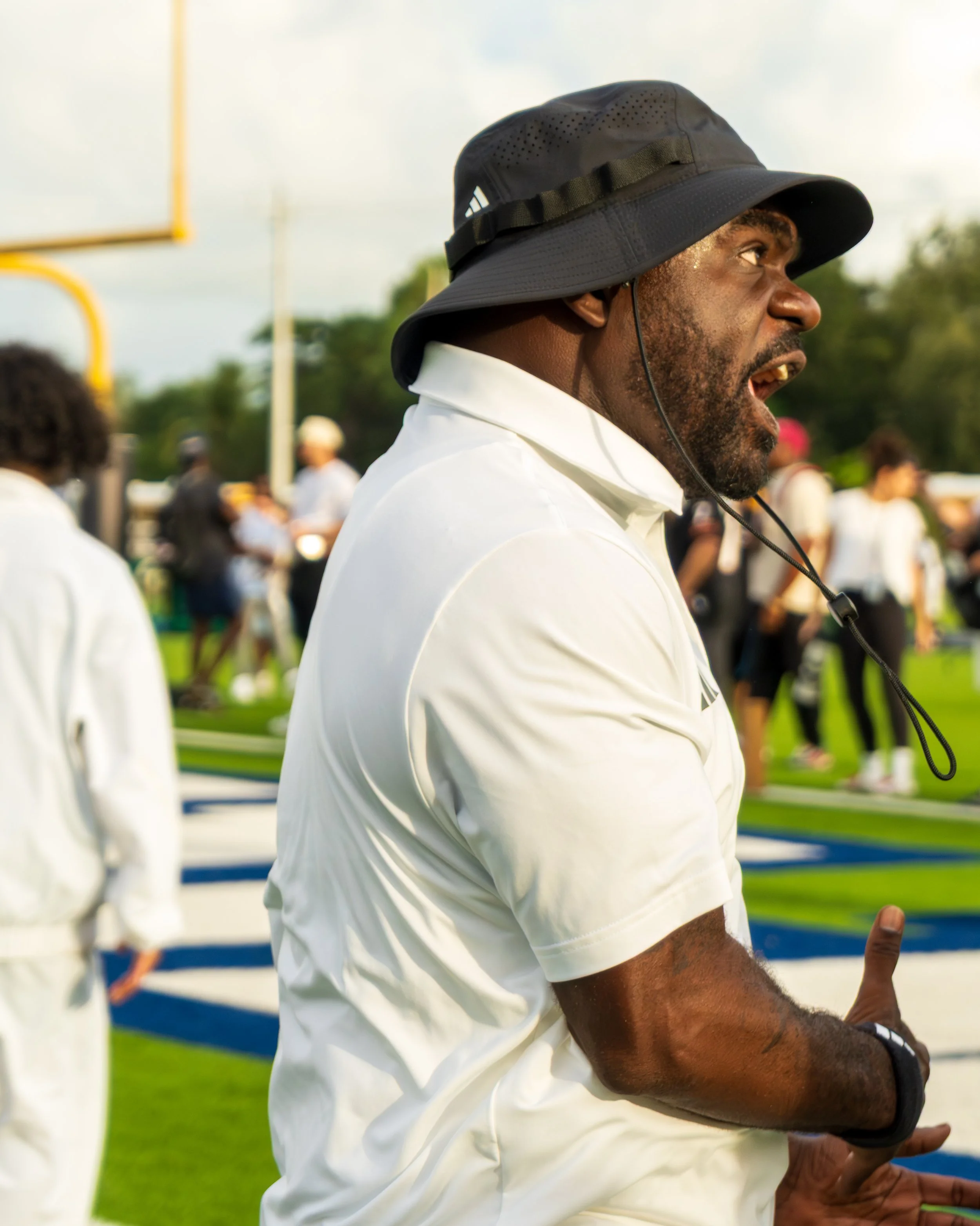 A man in a white shirt and a black bucket hat coaching or instructing, with a football field and people in the background.
