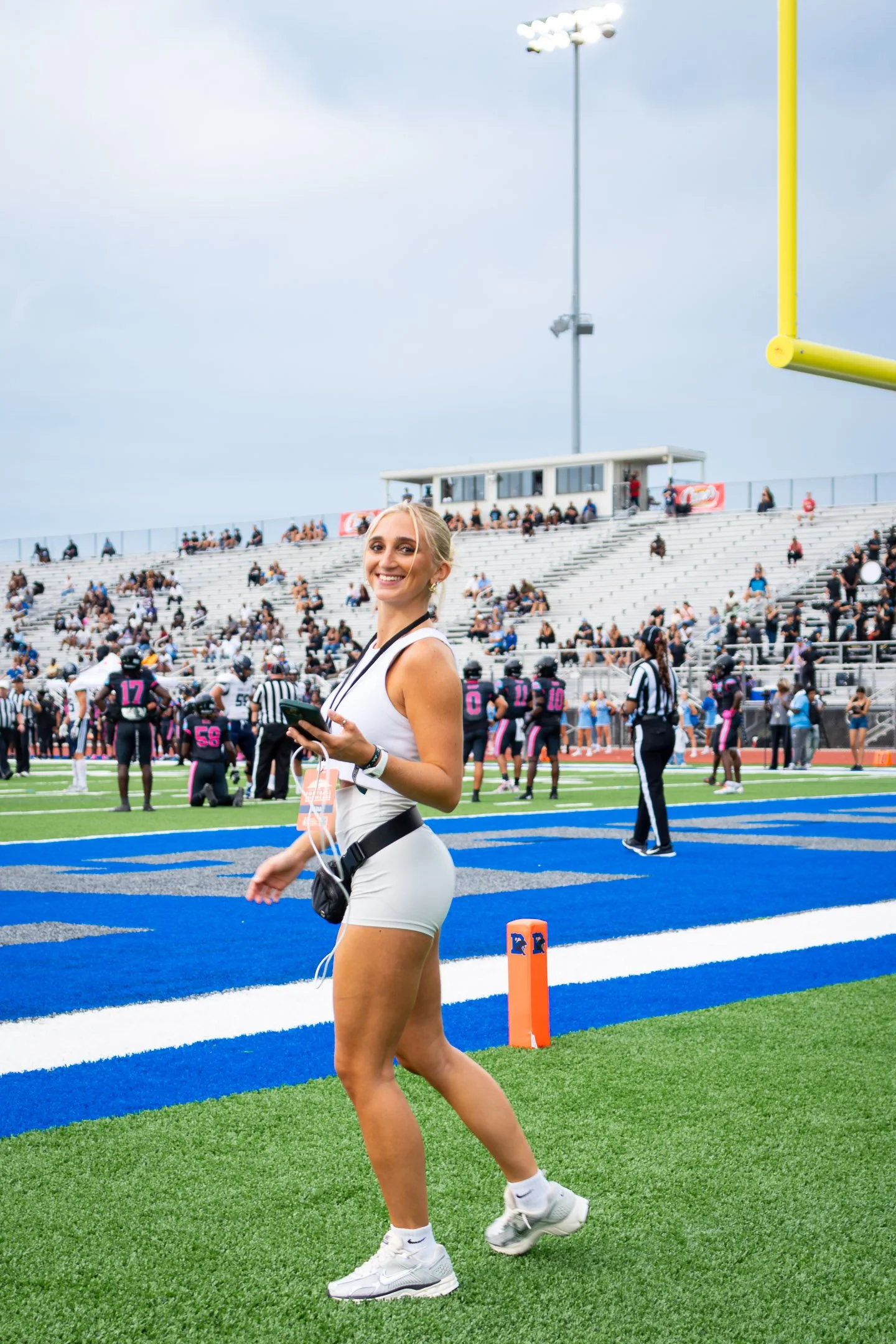 A smiling Ava Surinski in athletic attire walking along the sideline of a football field, with players and spectators in the background, during a game day.