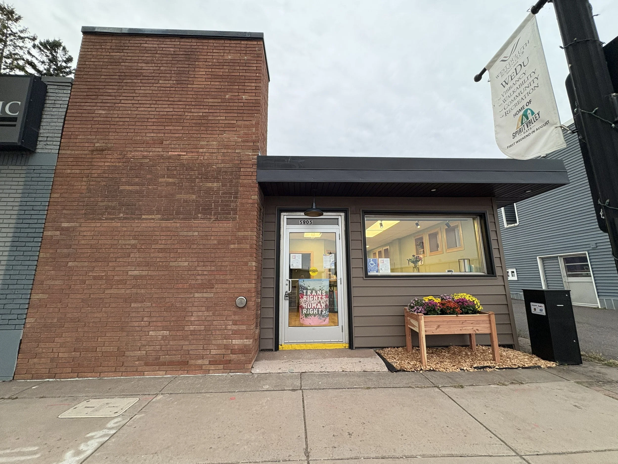 photograph of the pleasure and belonging storefront with a tall brick section next to a one story building with brown siding and a dark roof. The glass door and front window are lit from inside and PRIDE flags are visible through the glass.