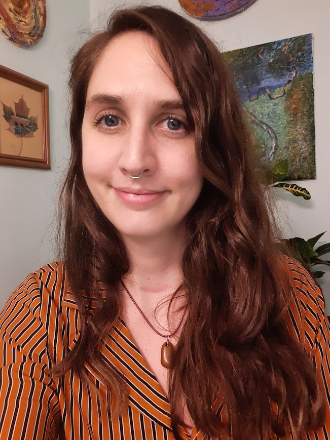 photo of Juniper a light-skinned non-binary person with long brown hair and a septum piercing smiling at the camera with artwork hanging behind them