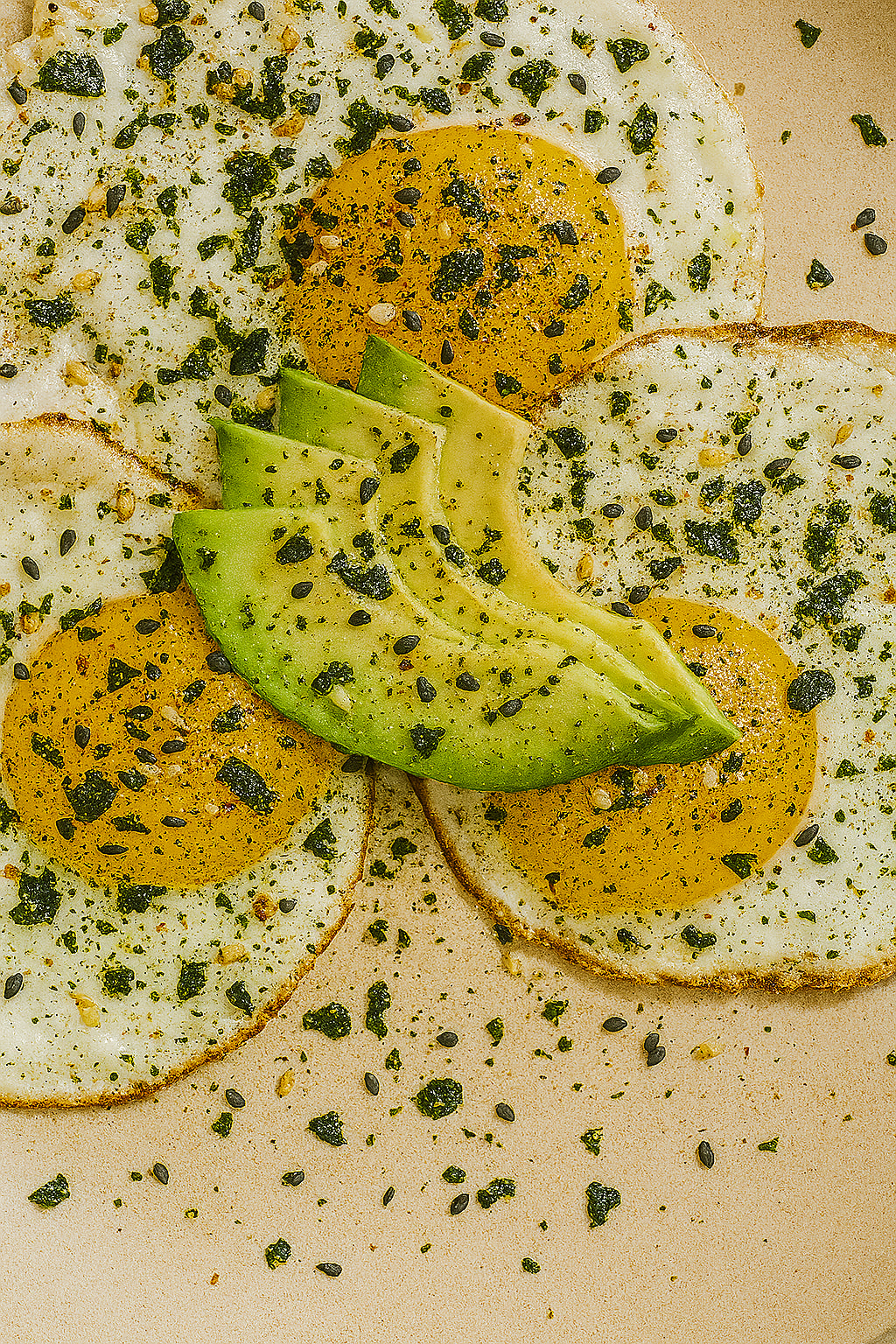 Close-up of three fried eggs with seasoning, sliced avocado, and black pepper, on a beige surface.