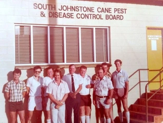 Group of nine people standing outside the South Johnstone Cane Pest & Disease Control Board building, which has red shutters and a staircase with yellow handrails.