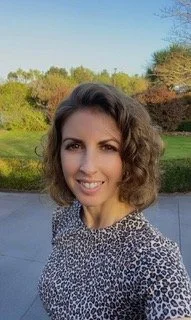 Jess, an audiologist with shoulder-length curly hair smiling outdoors on a sunny day with trees and a blue sky in the background.