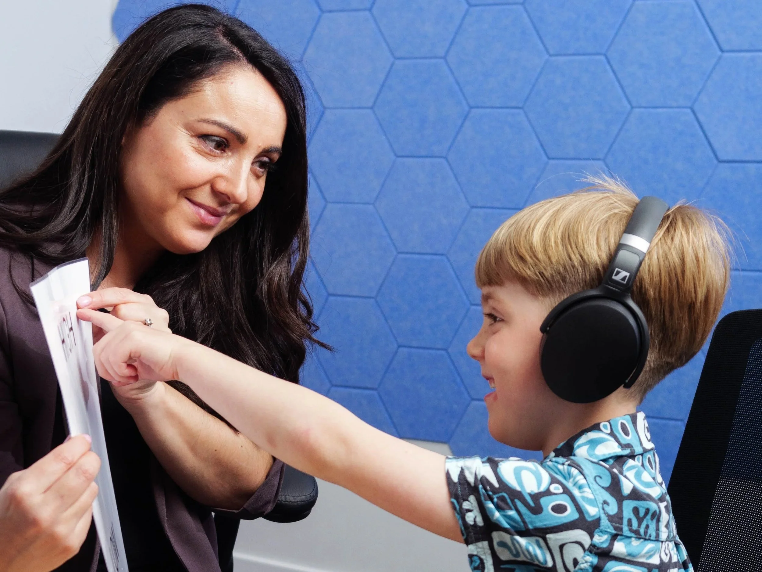 Katrin Hobson sitting at a desk and smiling as a young boy wearing headphones and a colourful shirt points to a book during a hearing examination.