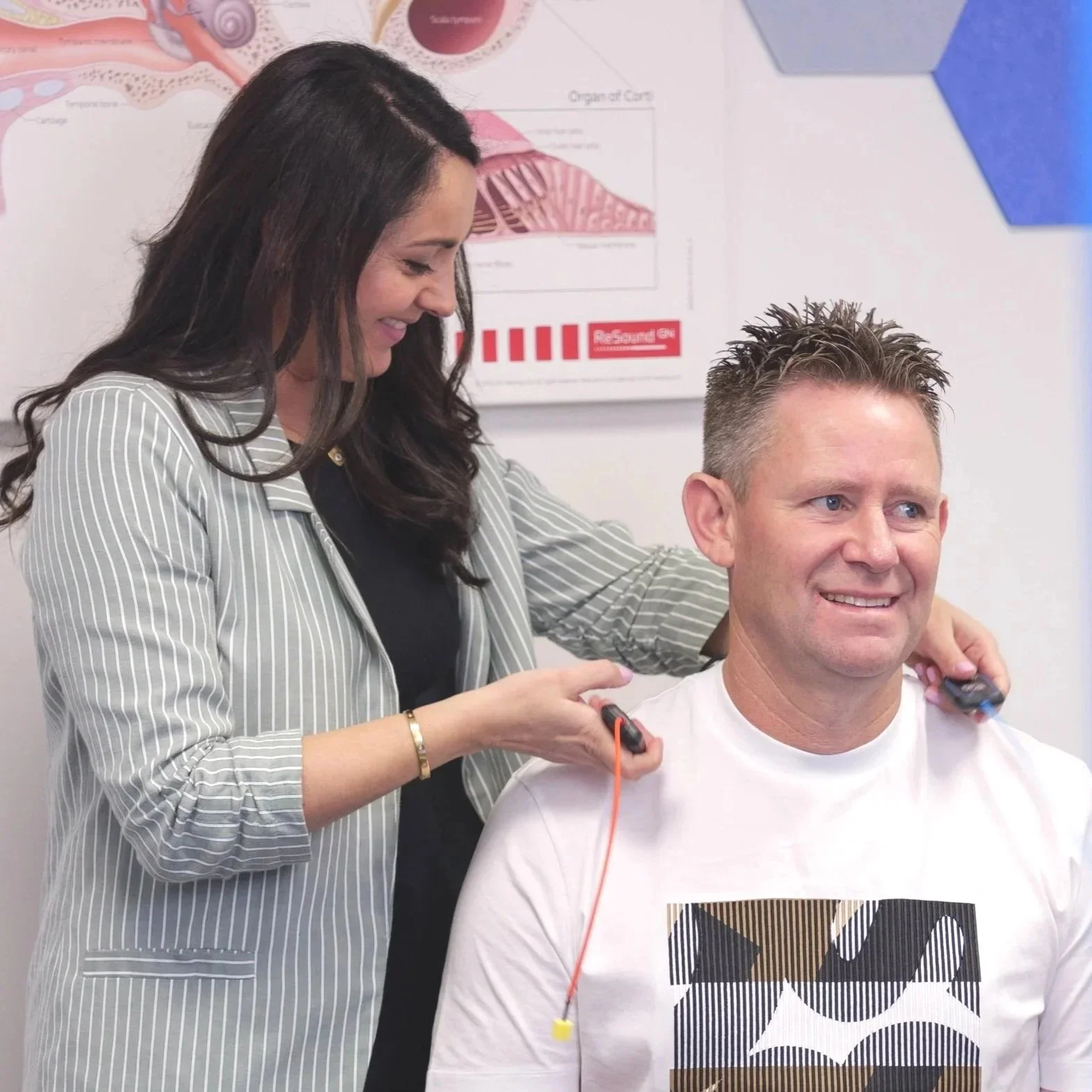Katrin Hobson, smiling as she uses an electrical device to test a man with short spiky hair, wearing a white graphic T-shirt's hearing, in an audiology clinic.