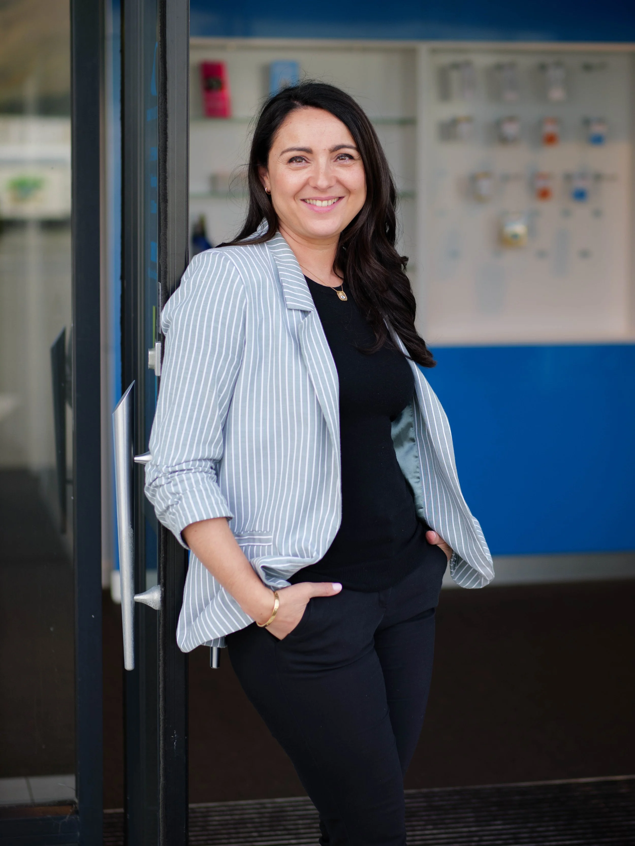 Katrin Hobson, smiling, with dark hair wearing a striped blazer, black top, and black pants, standing at the entrance of a building with her hands in her pockets.