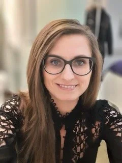 Bernadett, an Audiologist with long brown hair wearing glasses and a black lace top, smiling at the camera.